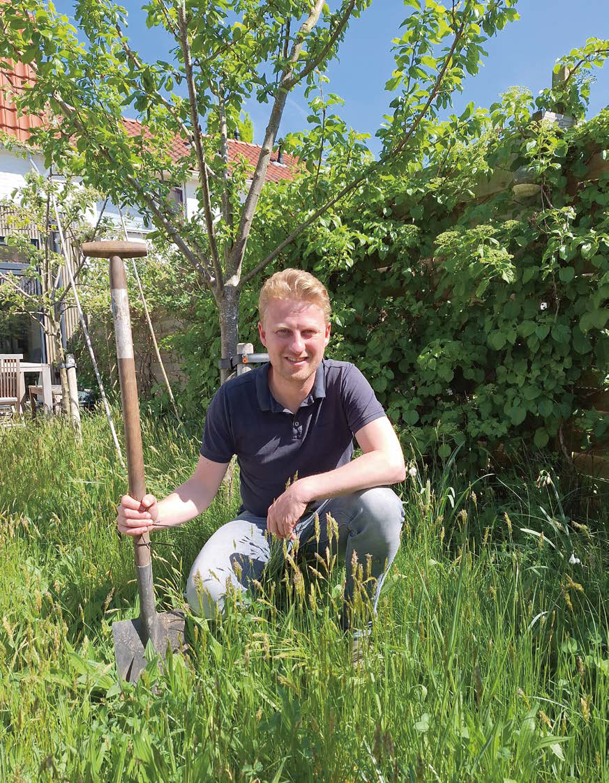 Man knielt in groene tuin met schep, omgeven door bomen en gras; huis op achtergrond.