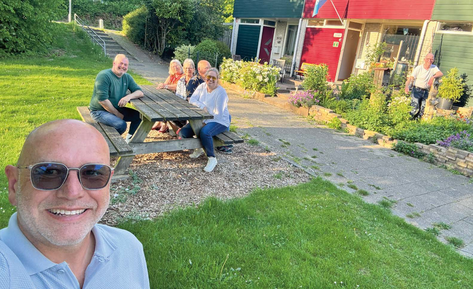 Een groep mensen zit buiten rond een picknicktafel op een zonnige dag. Een man maakt een selfie.