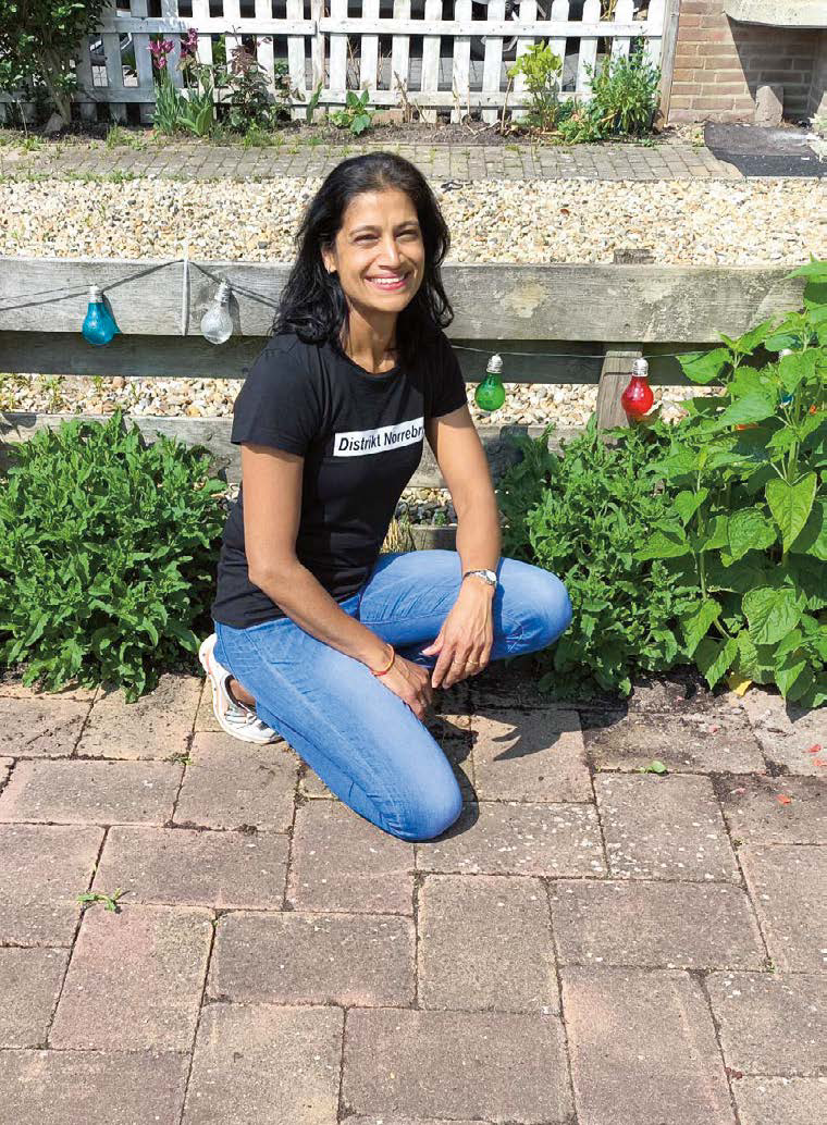 Vrouw knielt op terras naast groene planten, draagt een zwart T-shirt en blauwe jeans, glimlacht.
