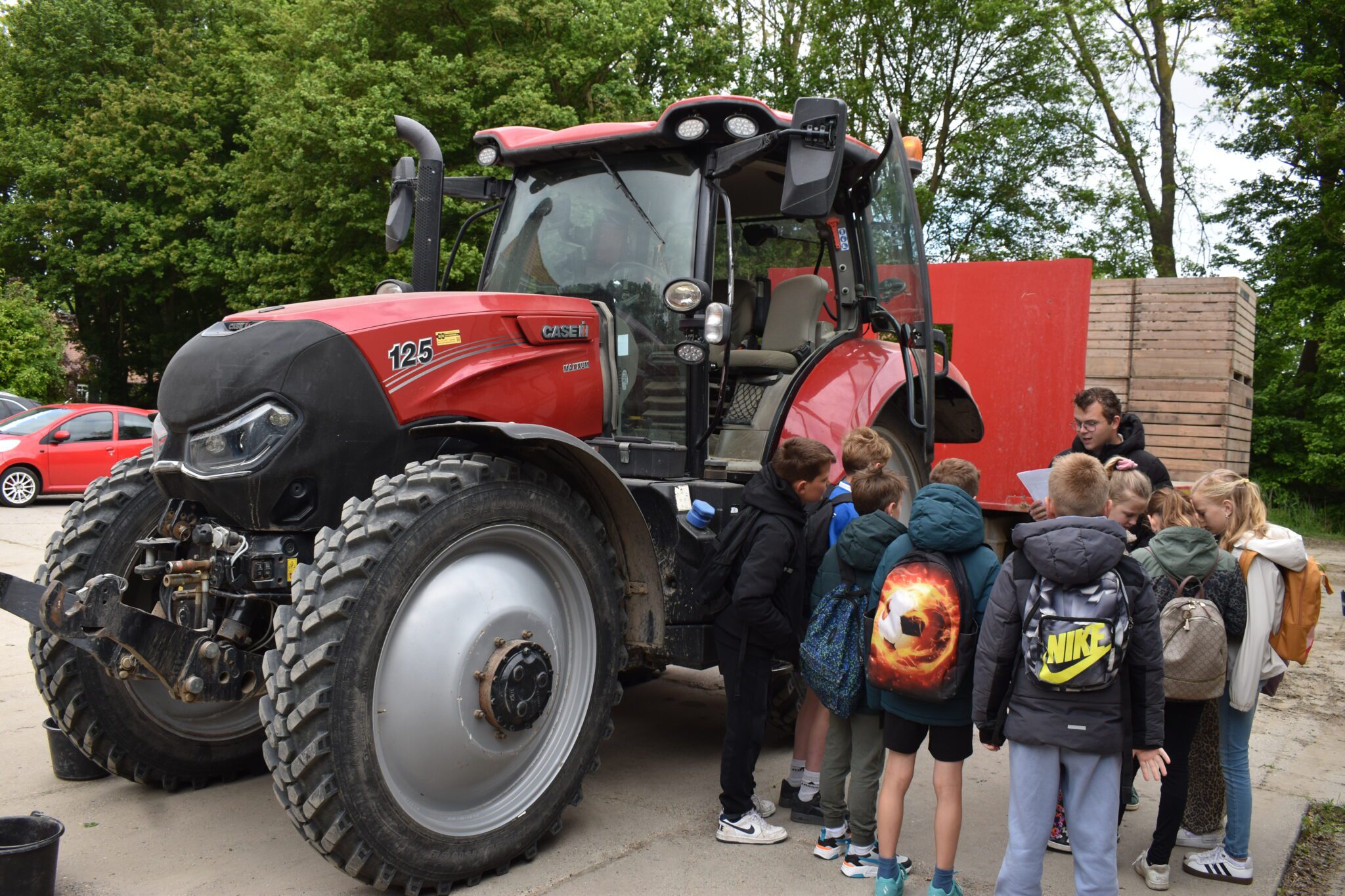 Kinderen luisteren aandachtig naar uitleg bij een grote rode tractor op een buitenlocatie.