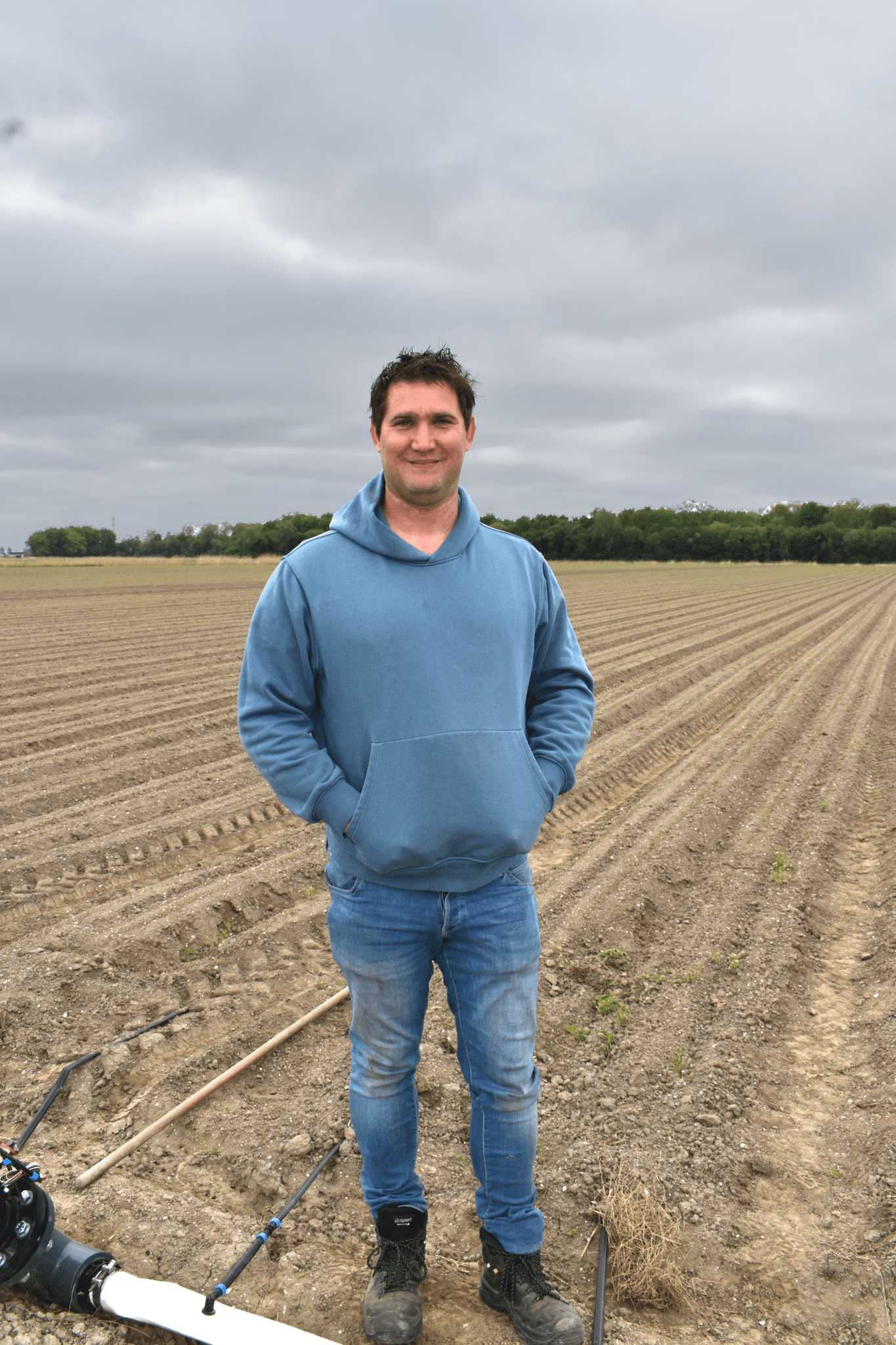 Man in blauwe trui staat op een akker met bewolkte lucht, omringd door gereedschap en landbouwgrond.
