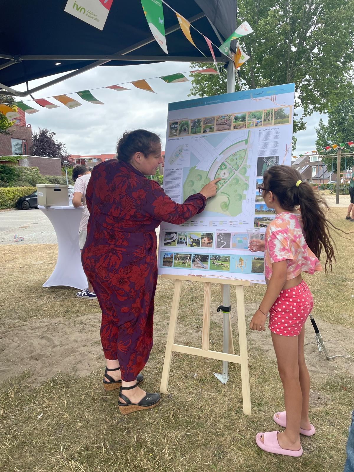 Een vrouw wijst op een groenkaartenbord, naast een kind, op een buitenbijeenkomst.