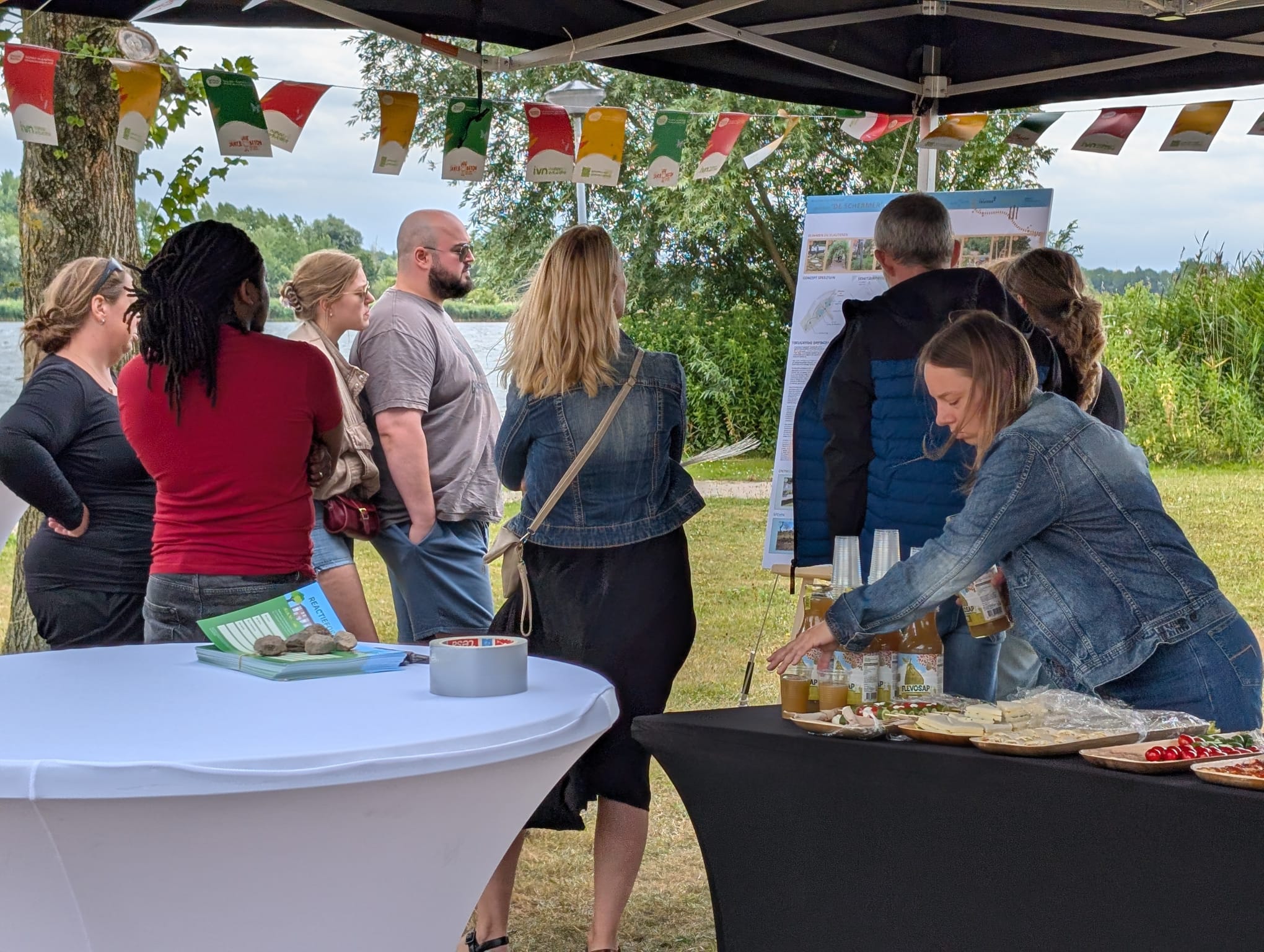 Mensen verzamelen onder een tent bij een voedselstand en informatiebord in een park.