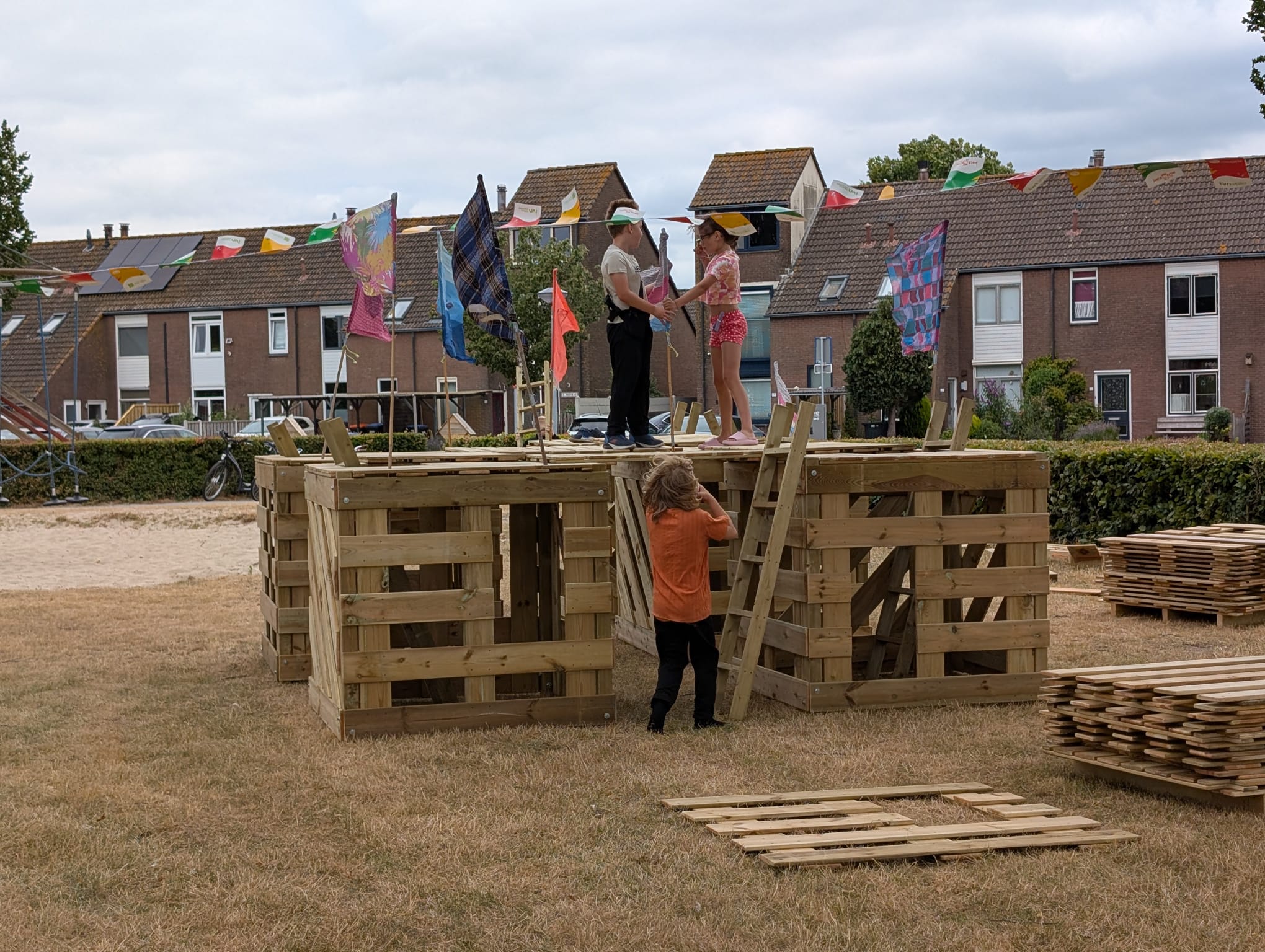 Kinderen spelen op een zelfgemaakt houten fort met vlaggen, in een grasveld bij rijtjeshuizen.