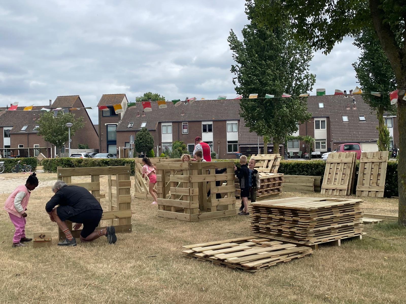 Mensen bouwen houten structuren met pallets op een grasveld in een woonwijk.