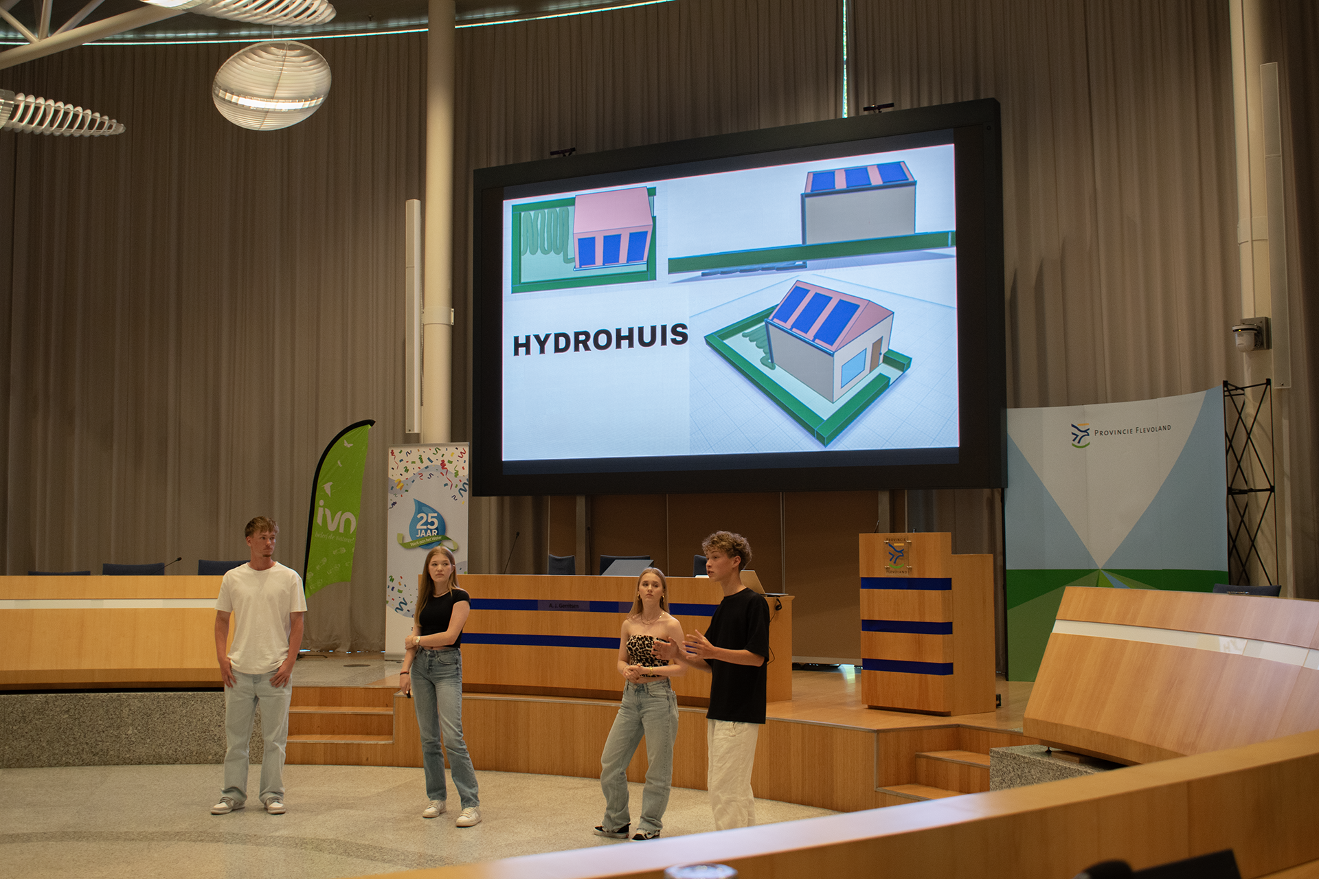Vier mensen presenteren over een 'Hydrohuis' op een groot scherm in een conferentieruimte.