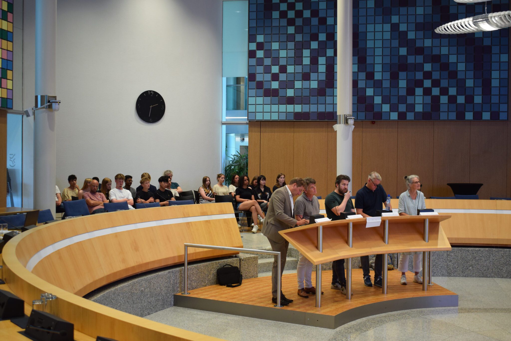 Mensen in een vergaderzaal, zittend en staand achter een spreekgestoelte.