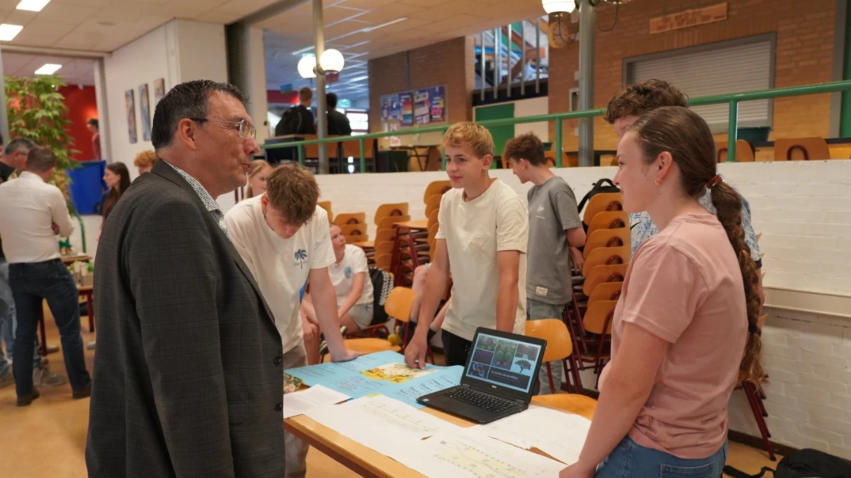 Mensen bespreken een project rond een tafel met een laptop en papieren in een schoolomgeving.