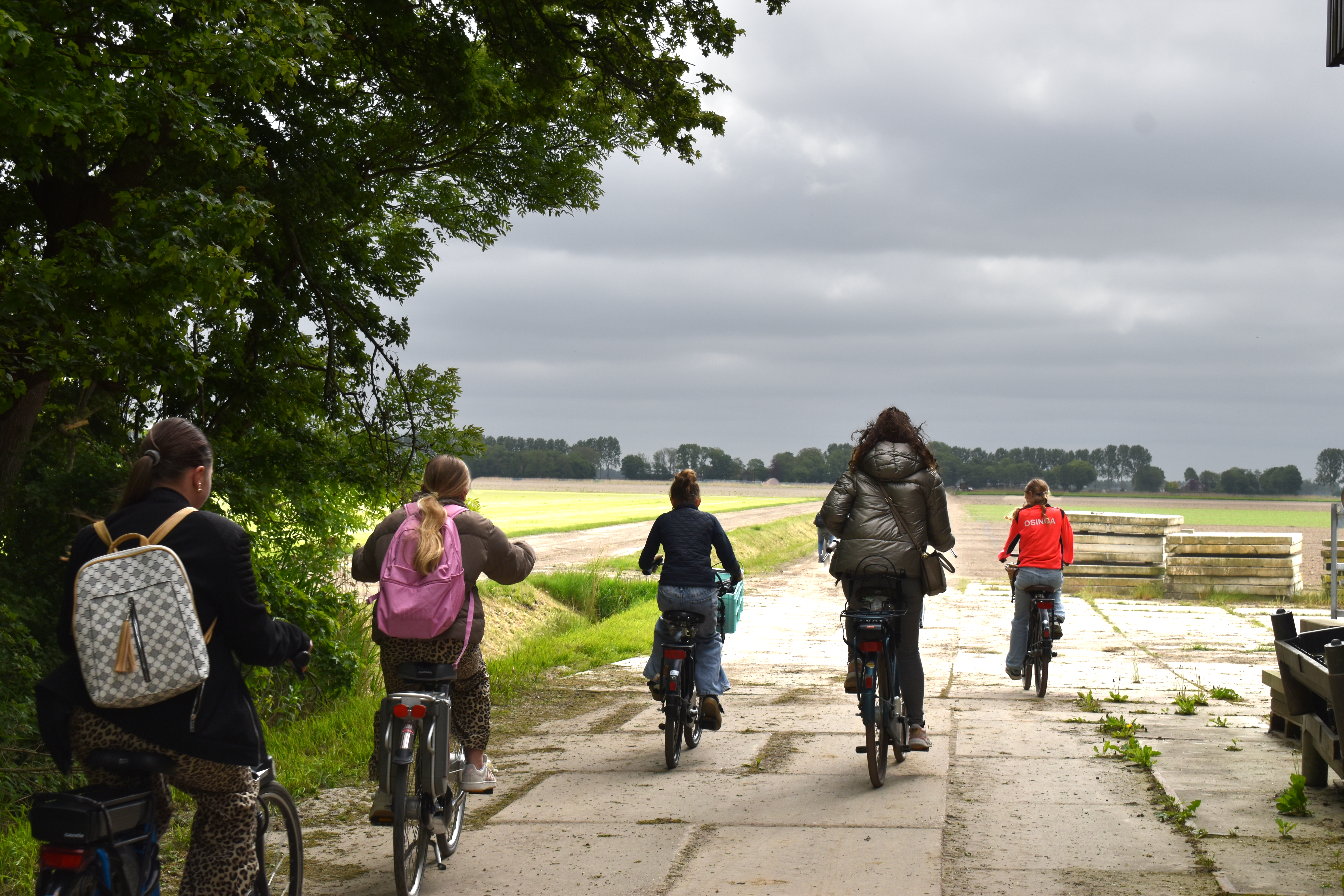 Groep fietsers op een plattelandsweg omringd door bomen en velden onder bewolkte lucht.