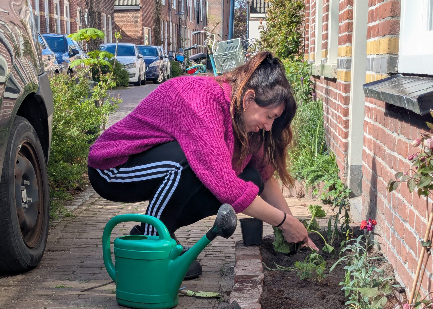Vrouw plant bloemen in een stadstuin, met een gieter naast haar op de stoep.