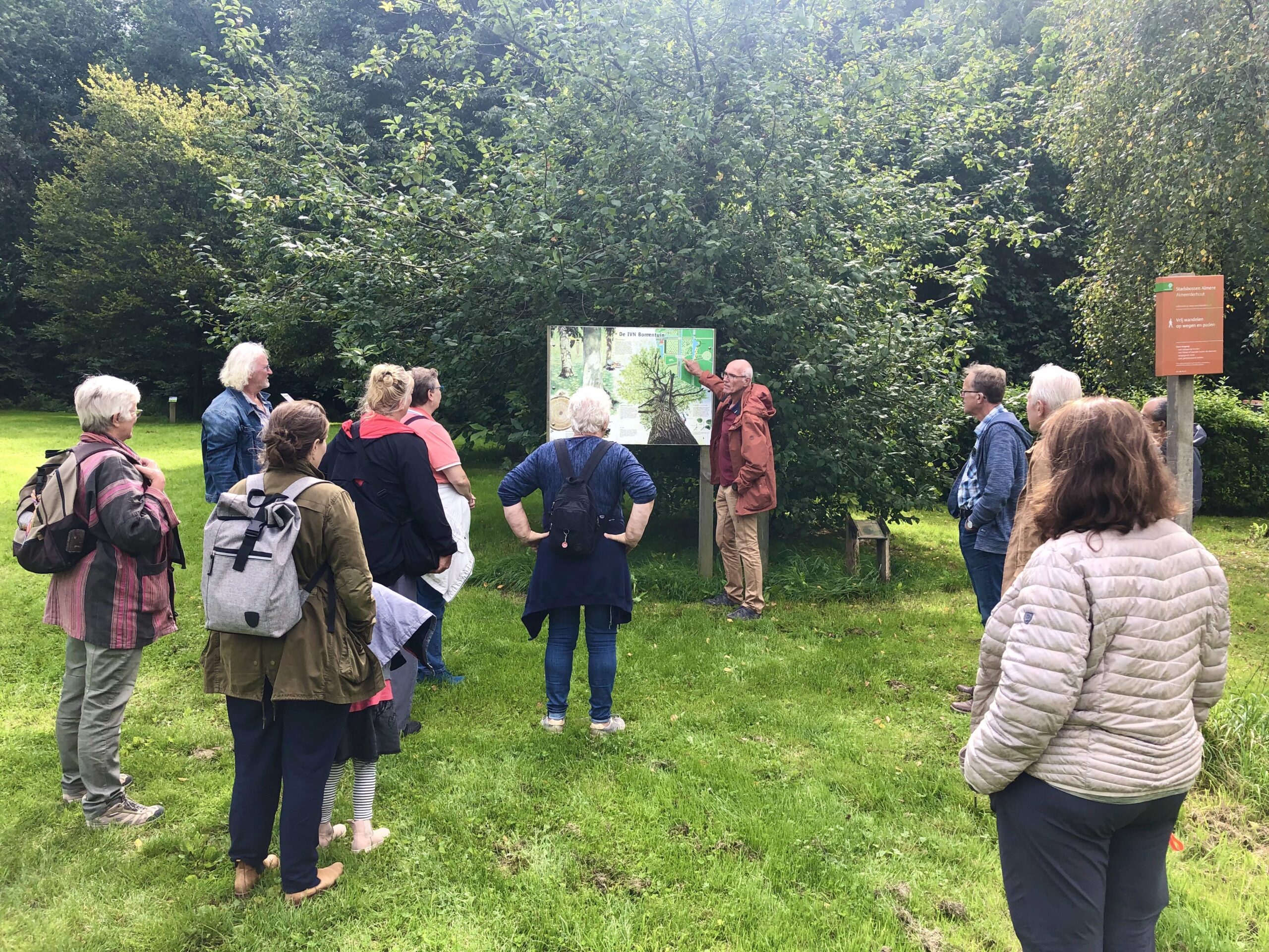 Een groep mensen luistert naar een gids bij een informatiebord in een groen park.