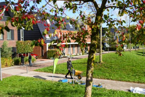 Een vrouw met een kruiwagen loopt langs moderne rijtjeshuizen en een groene, parkachtige omgeving.