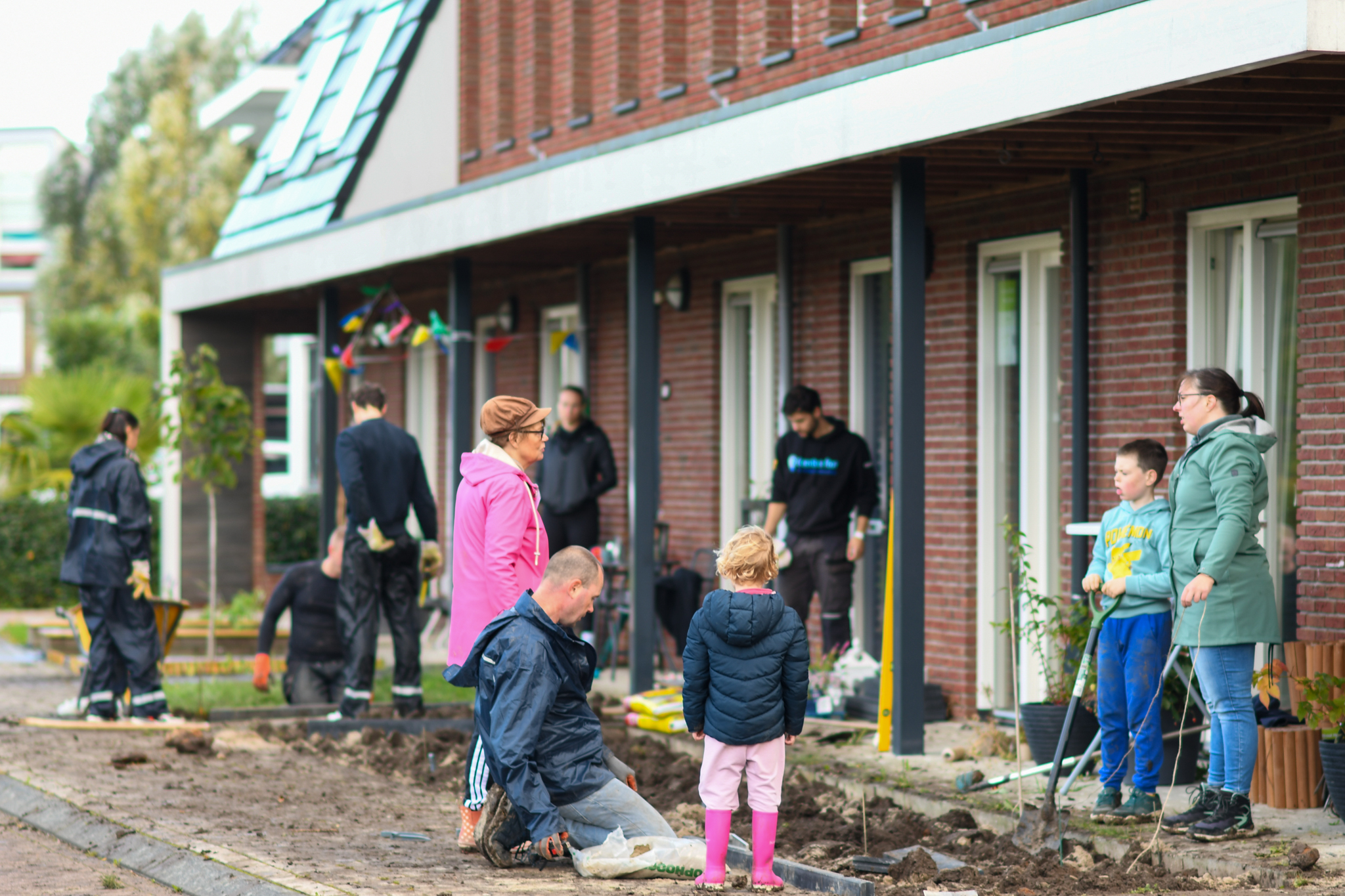 Mensen werken gezamenlijk aan tuinprojecten voor een rij huizen. Kinderen helpen mee.