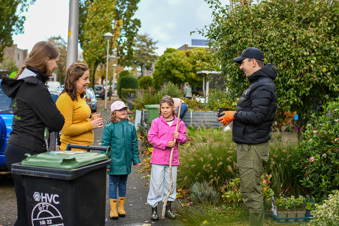 Mensen praten op een stoep naast een tuin, met kinderen in regenjassen en een man in een zwarte jas.