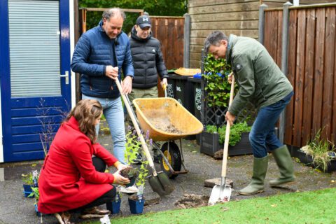 Vier mensen planten bloemen in een tuin met een kruiwagen op de achtergrond.