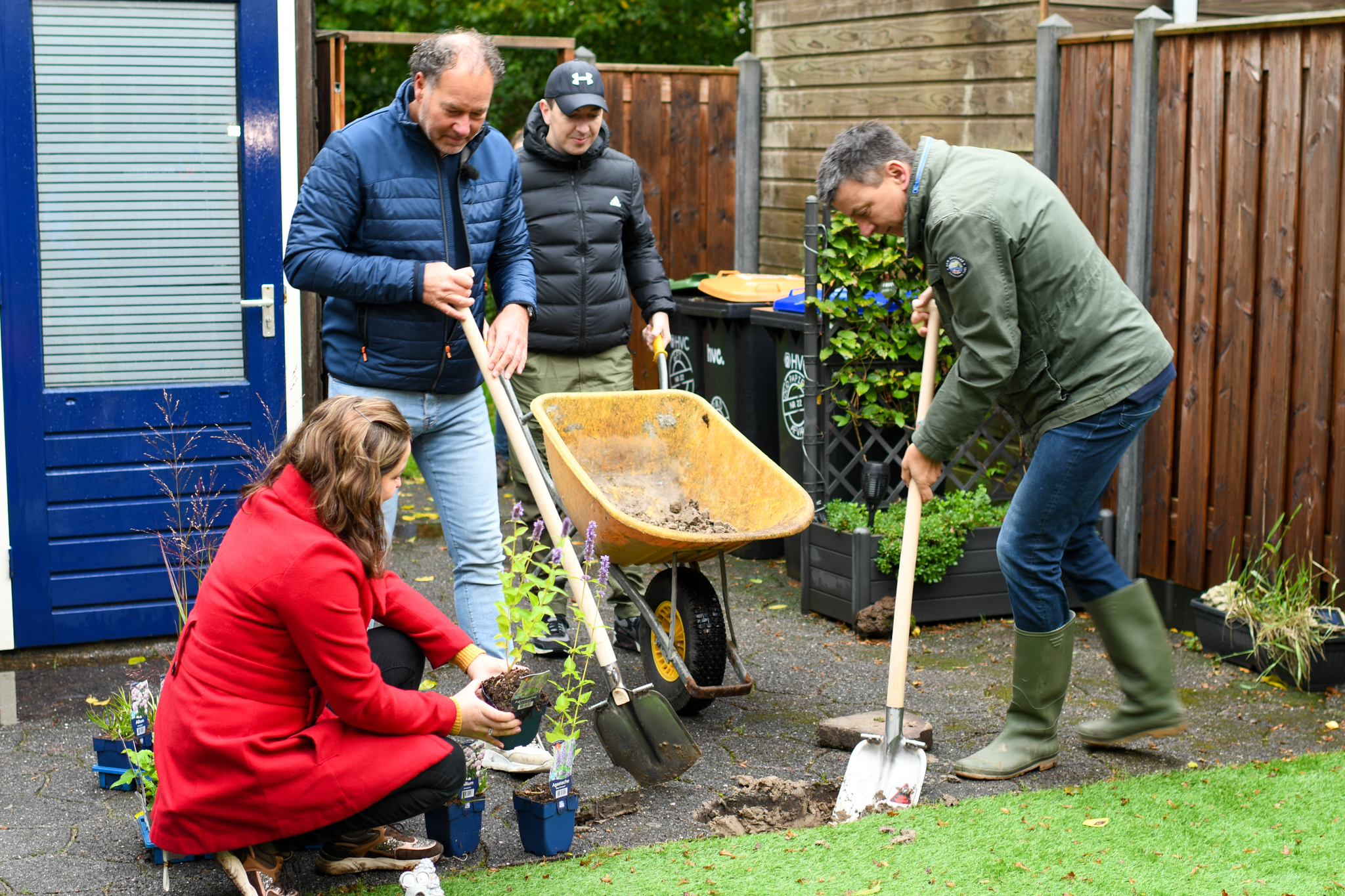 Vier mensen planten bloemen in een tuin met een kruiwagen op de achtergrond.