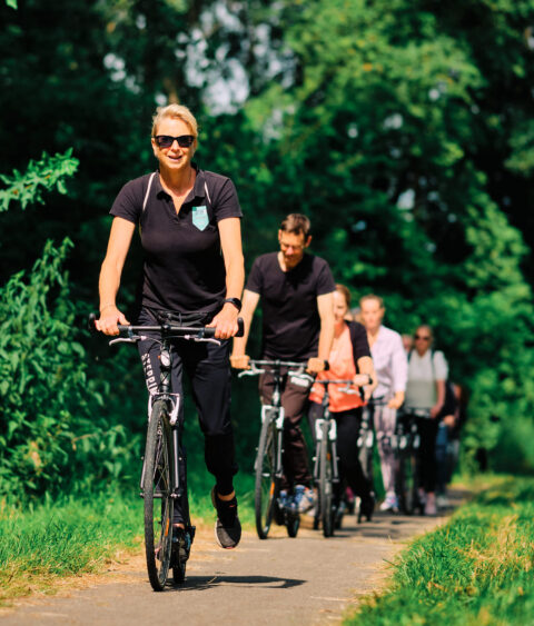 Groep mensen fietst op een pad, omlijst door groene bomen, met een vrouw op de voorgrond.