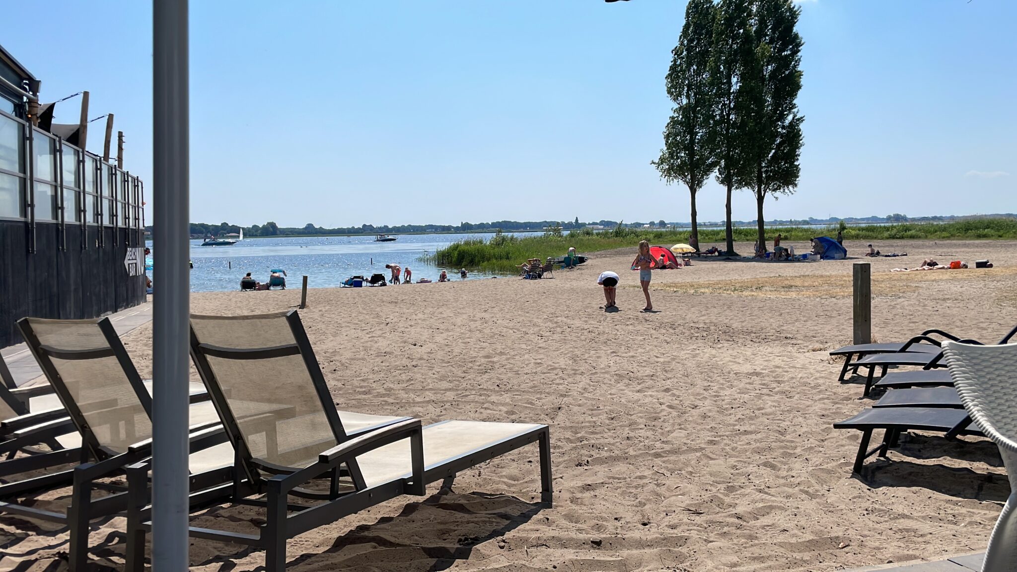 Strand met ligstoelen, mensen bij het water en bomen op de achtergrond onder een heldere hemel.