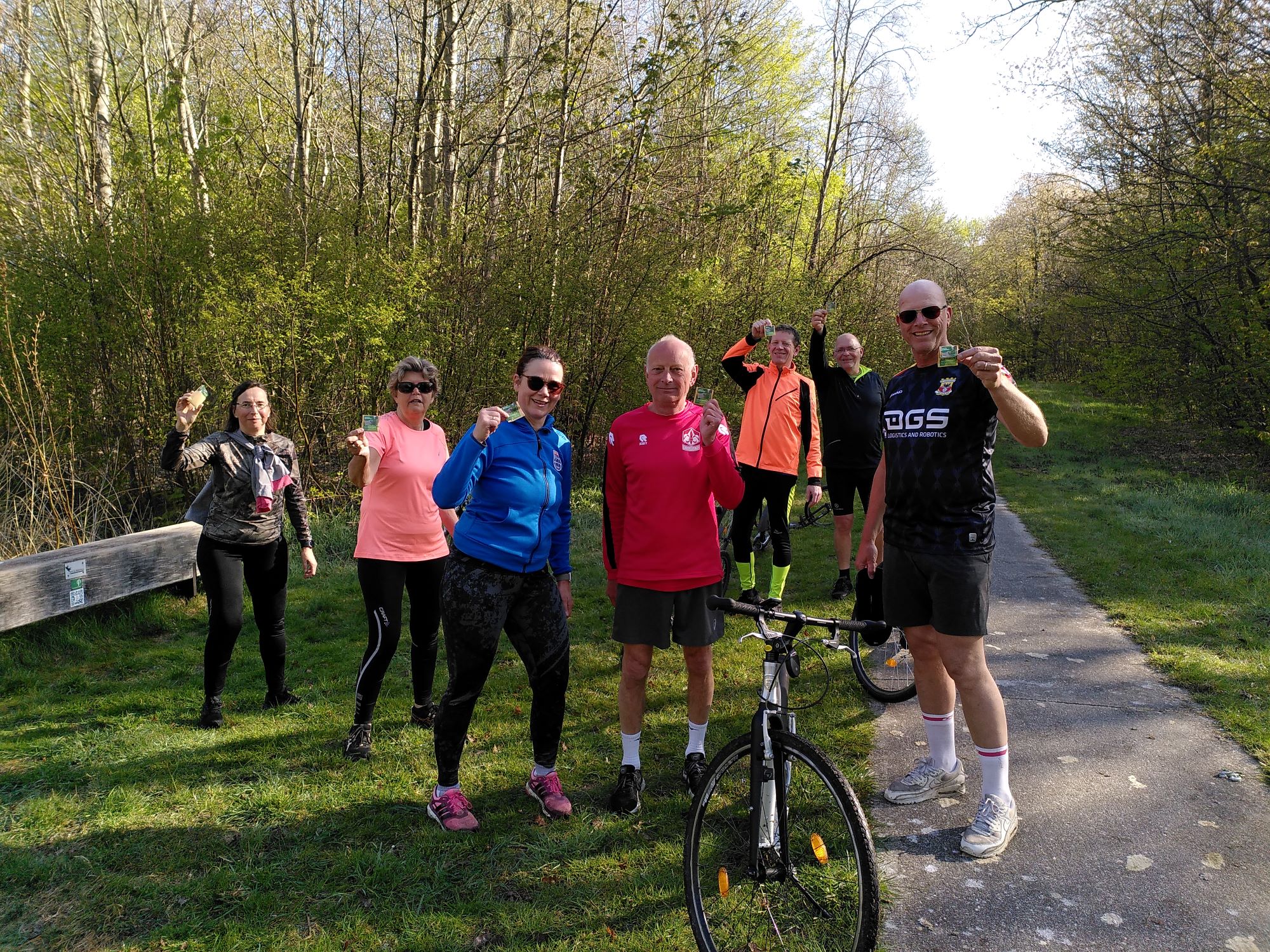 Groep mensen in sportkleding poseert met zakjes bloemzaad in een bosrijke omgeving.