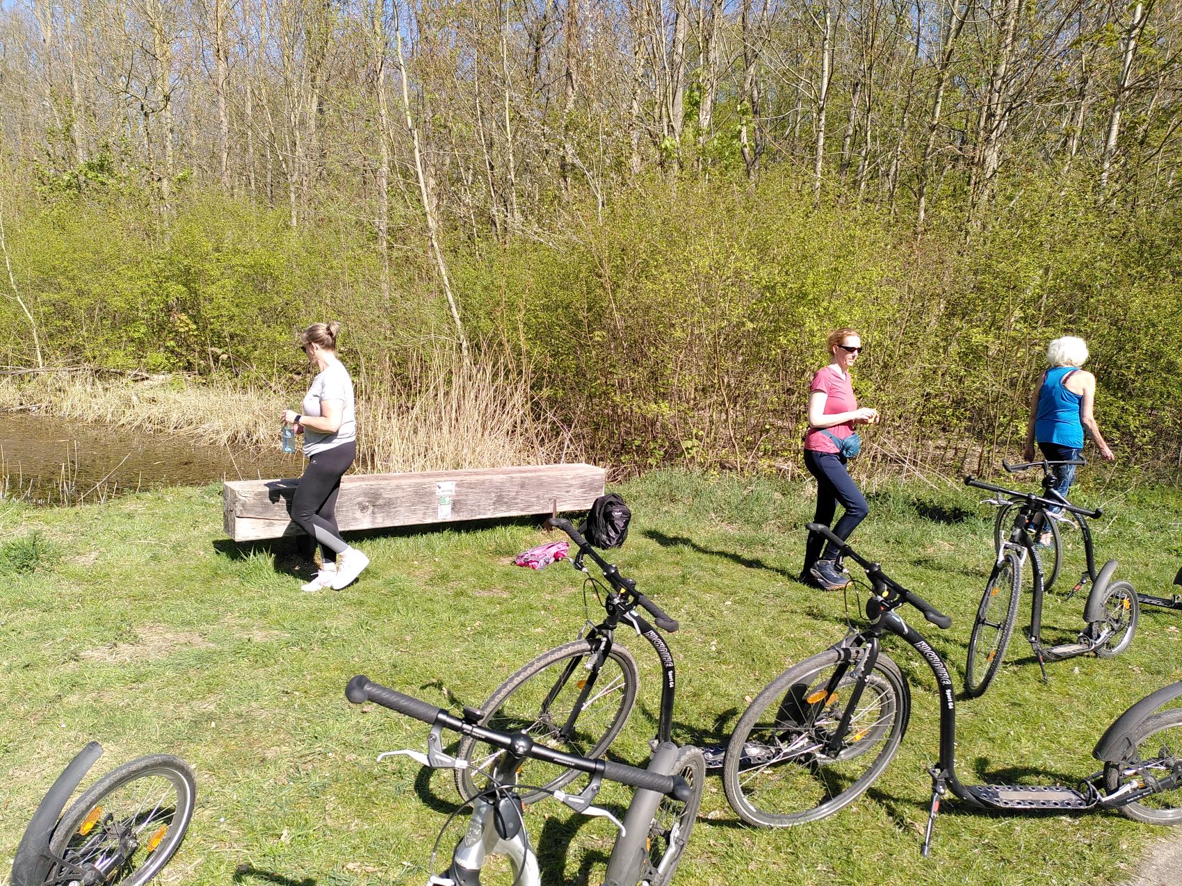 Een park met drie mensen in sportkleding, fietsen geparkeerd op het gras, bomen op de achtergrond.
