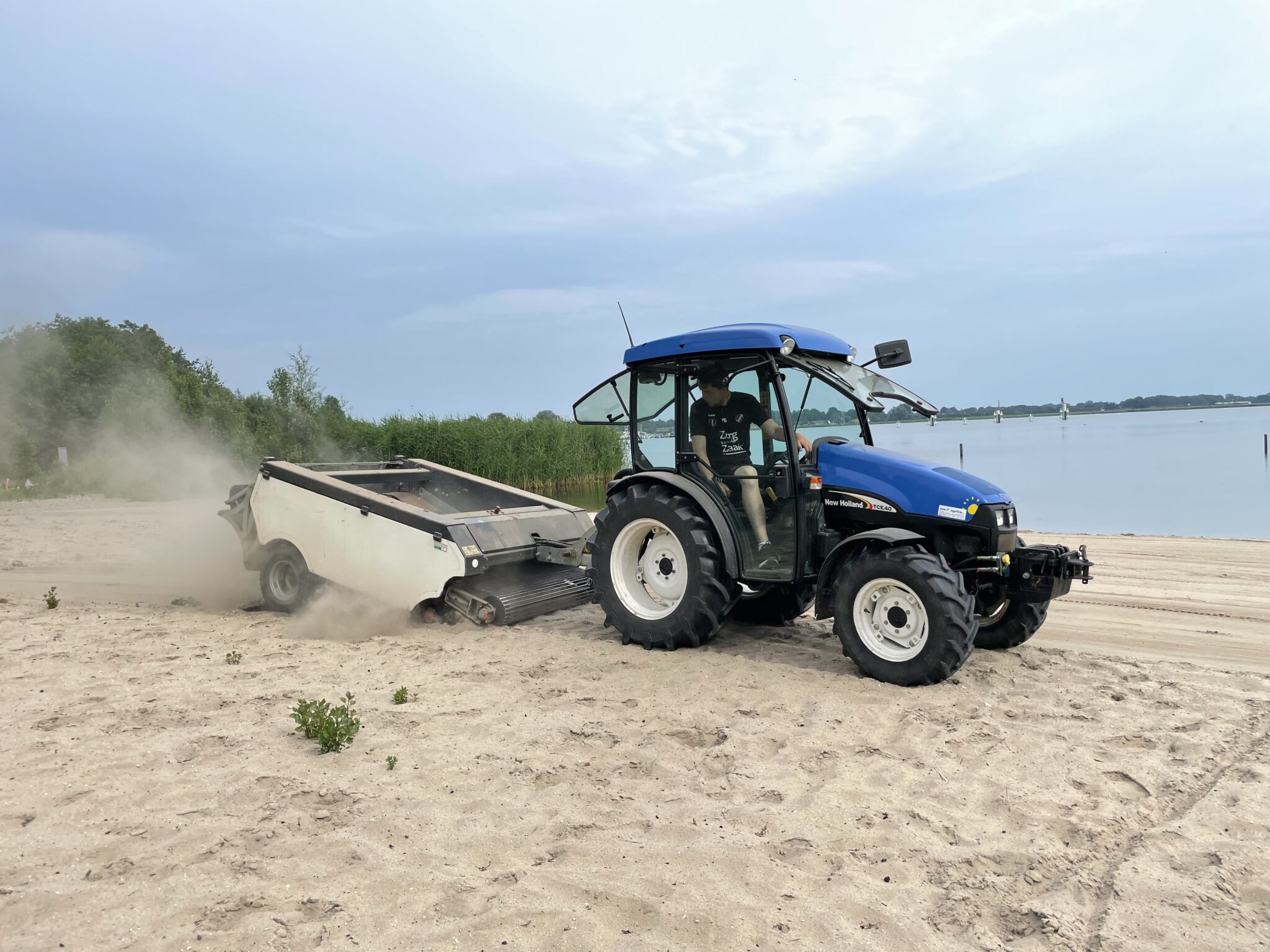 Tractor met aanhanger reinigt strand bij het water, bomen op de achtergrond.