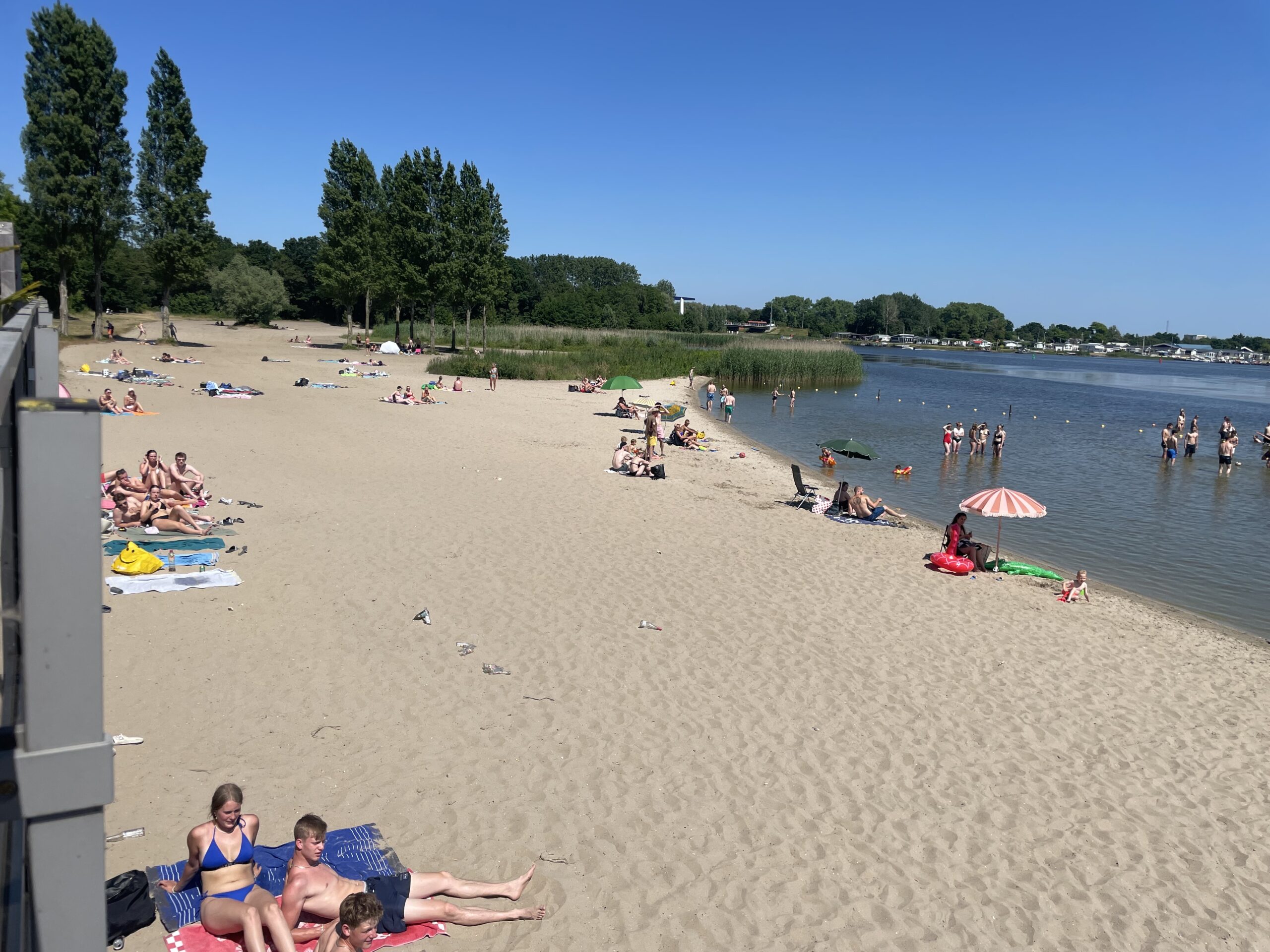 Strand vol mensen bij een meer, onder een heldere blauwe hemel.