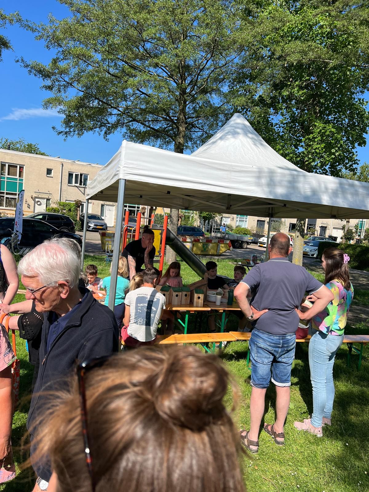 Mensen verzamelen onder een witte partytent in een park voor een buitenevenement op een zonnige dag.
