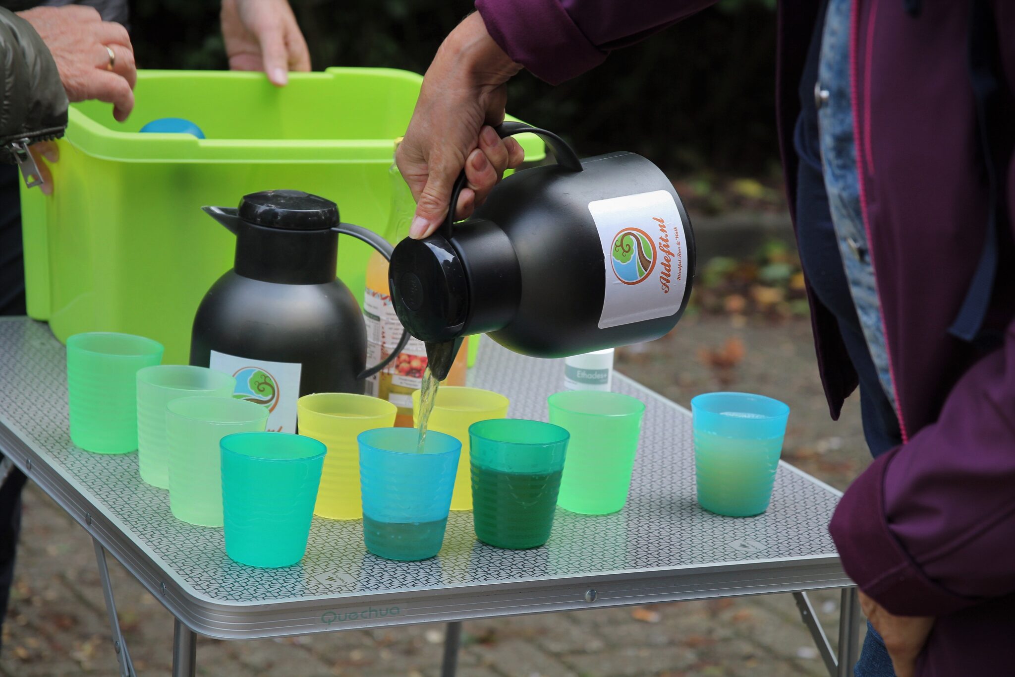 Persoon schenkt drank in gekleurde bekers op een tafel met groene bak.