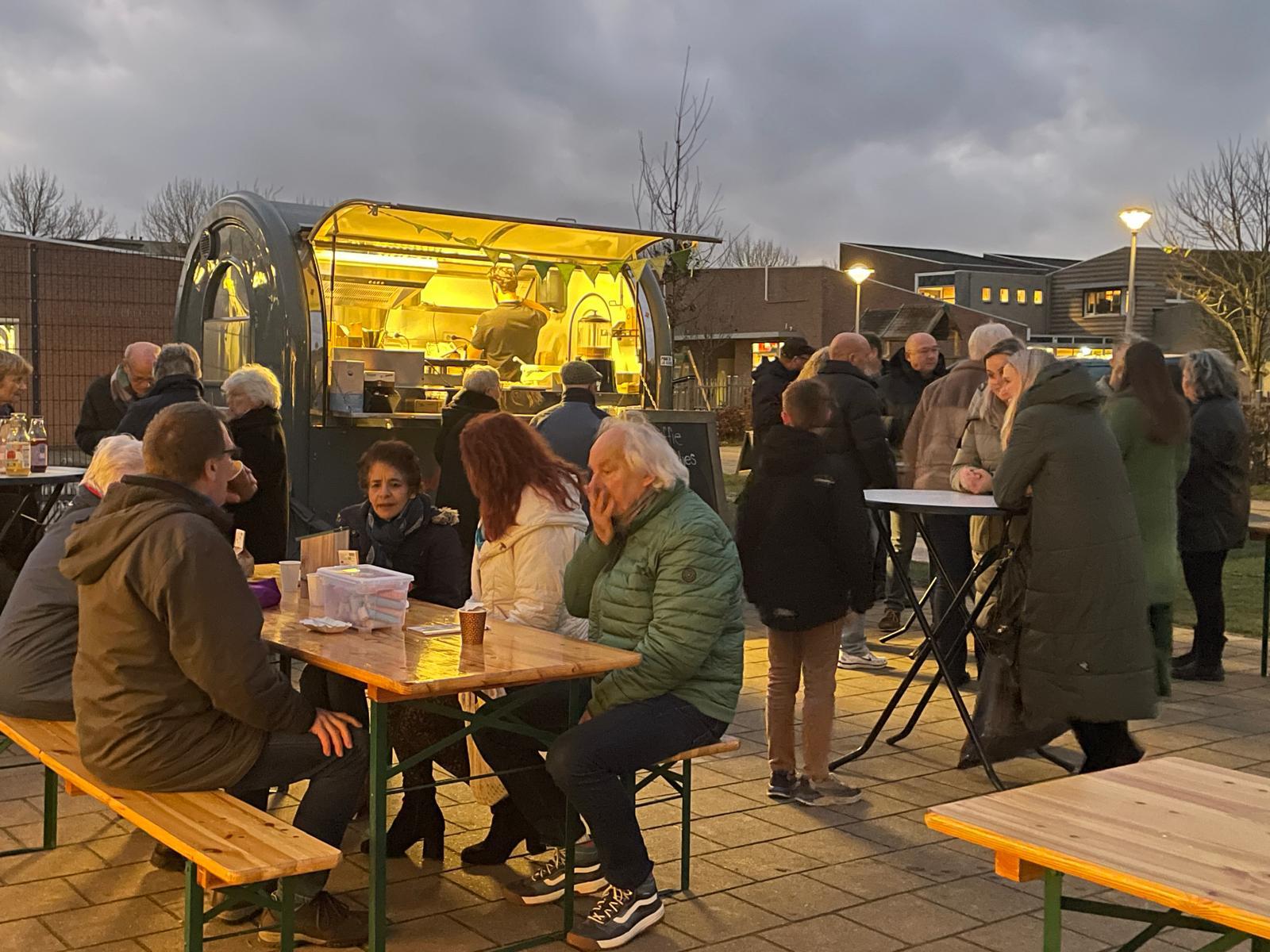 Groep mensen bij een foodtruck, zittend en staand, buiten in de schemering.