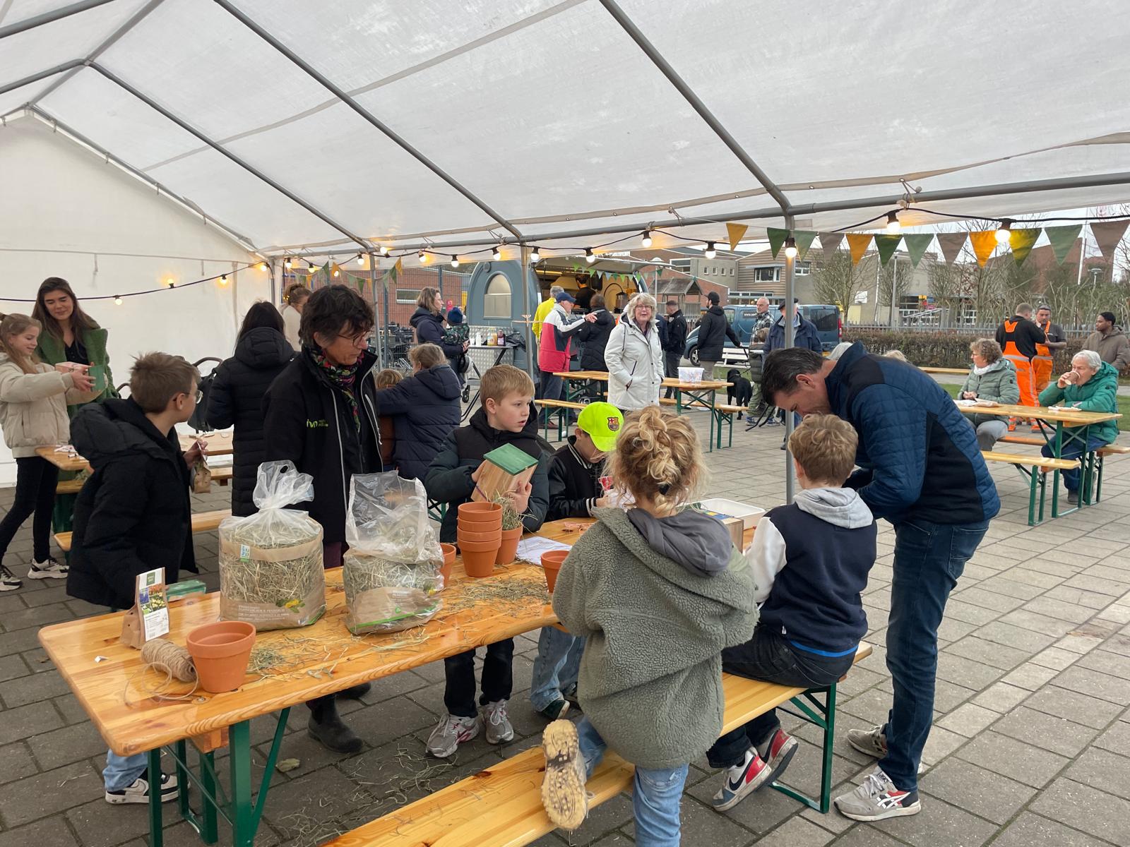 Mensen in een tent op een buitenmarkt, bezig met activiteiten aan houten tafels.