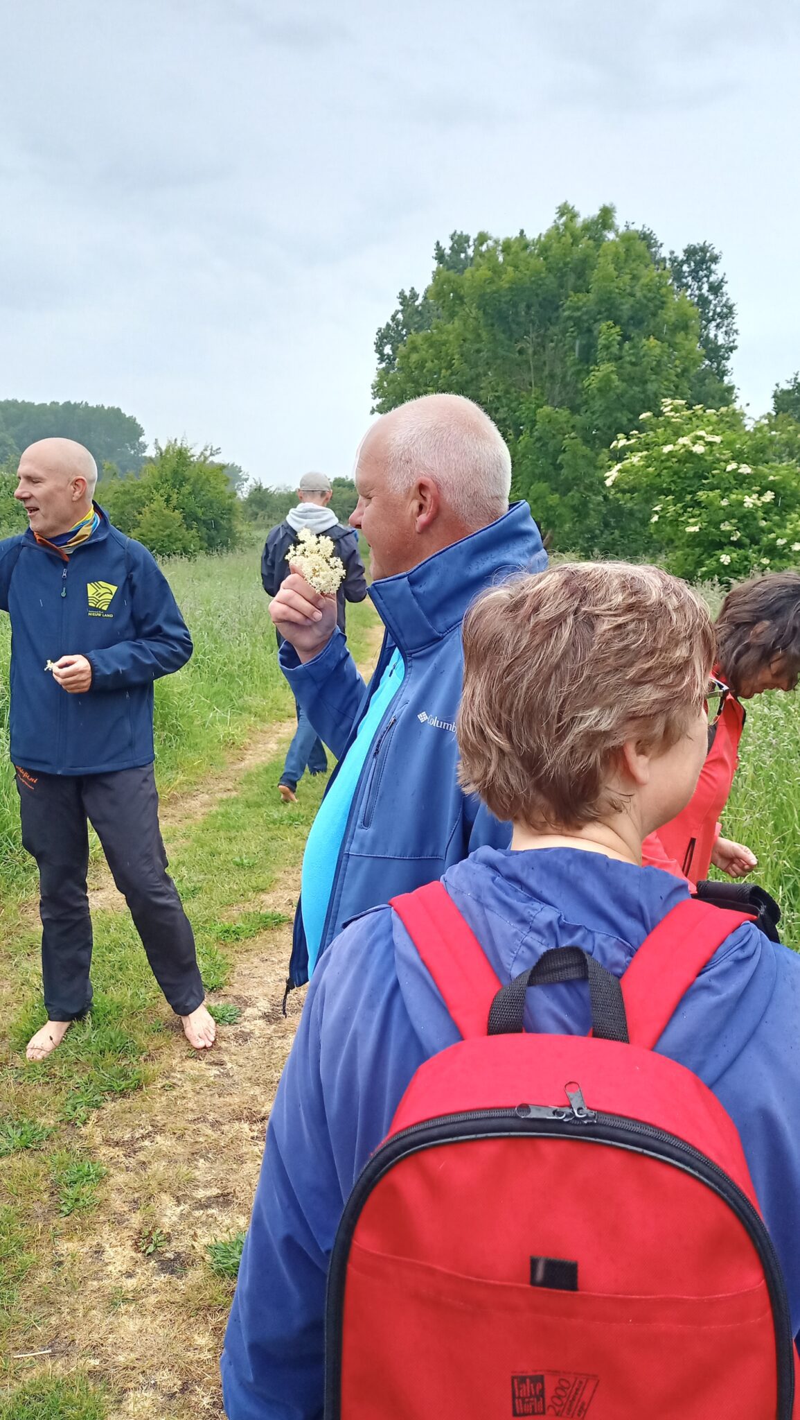 Mensen op een natuurpad, sommige bloesems vasthoudend, omgeven door groen en bomen.