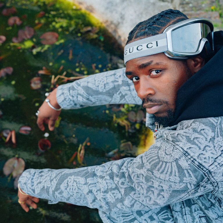 Man met gevlochten haar en Gucci-bril kijkt omhoog naast een vijver met lelies en mos.
