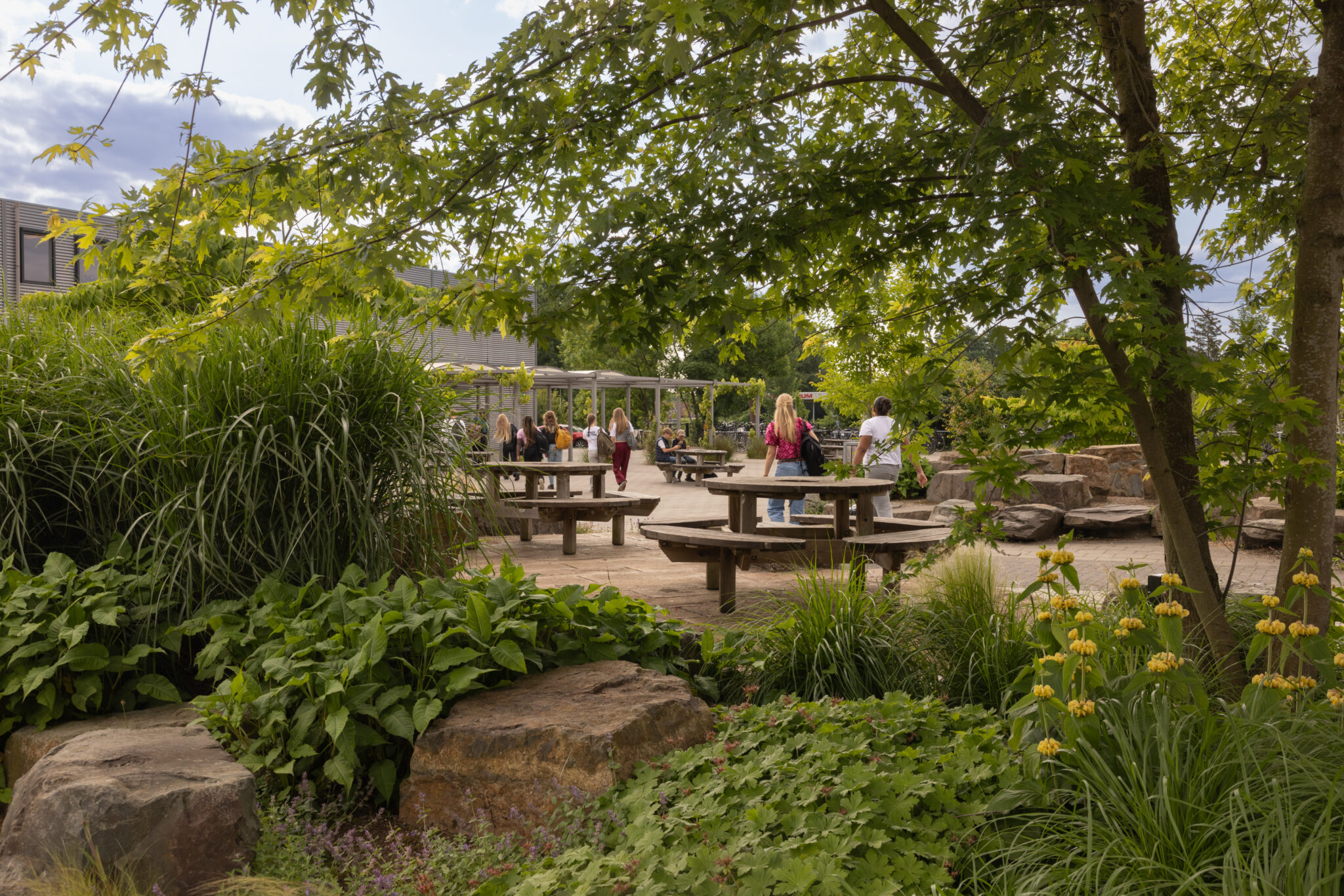 Mensen bij picknicktafels in een groene tuin met bomen en struiken, bij een zonnig terras.
