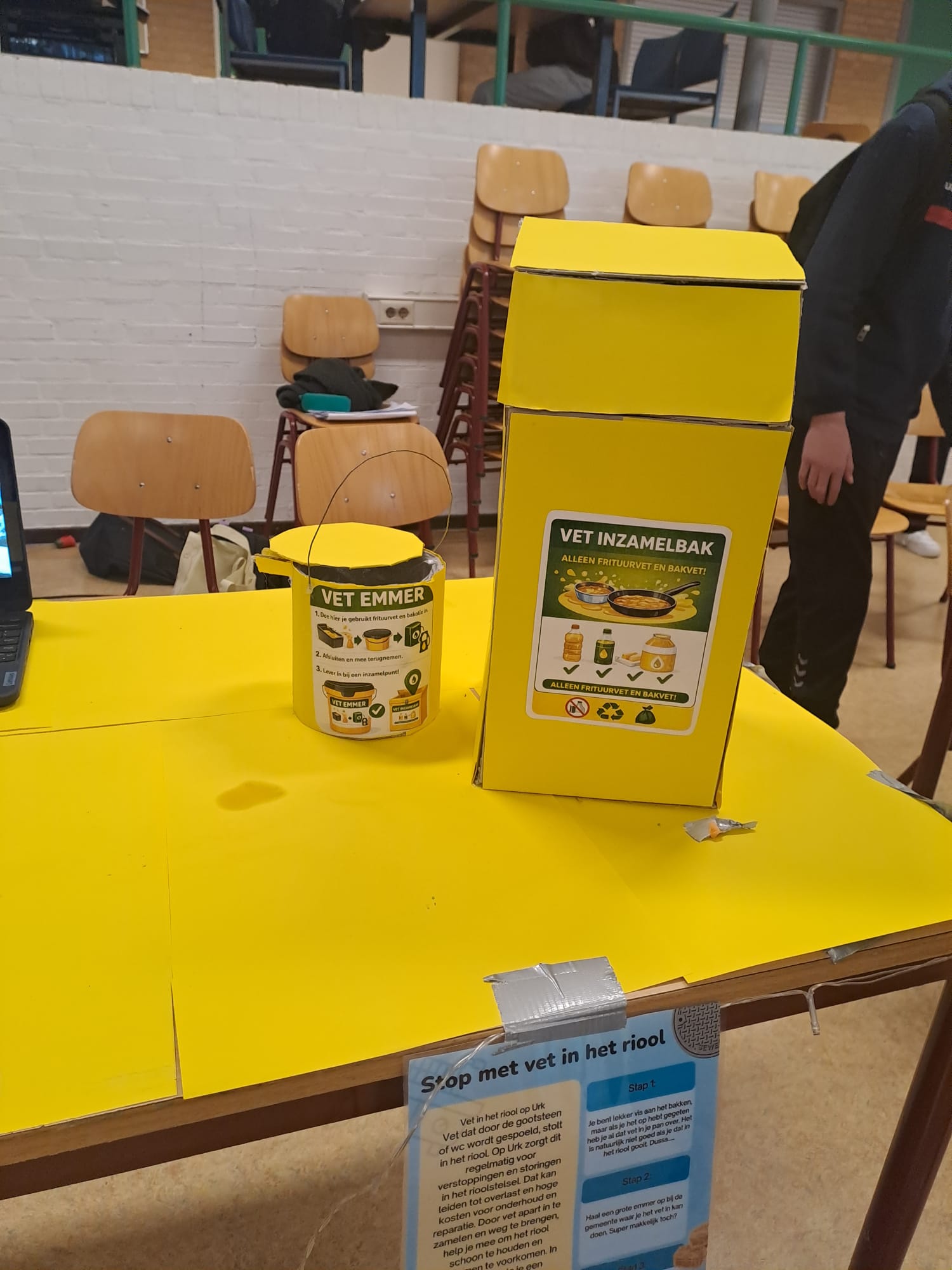 Gele vet inleverbak en emmer op tafel, met poster over vetafvalinzameling.