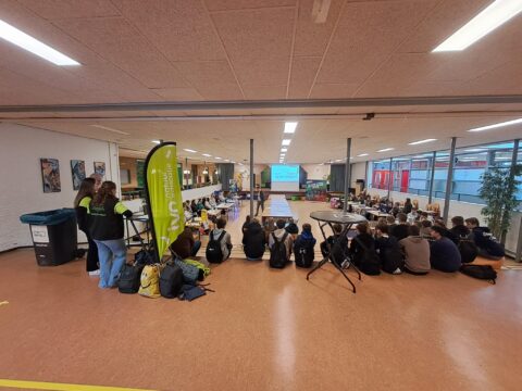 Groep mensen op een bijeenkomst in een hal; presentatiescherm en IVD-banner zichtbaar.