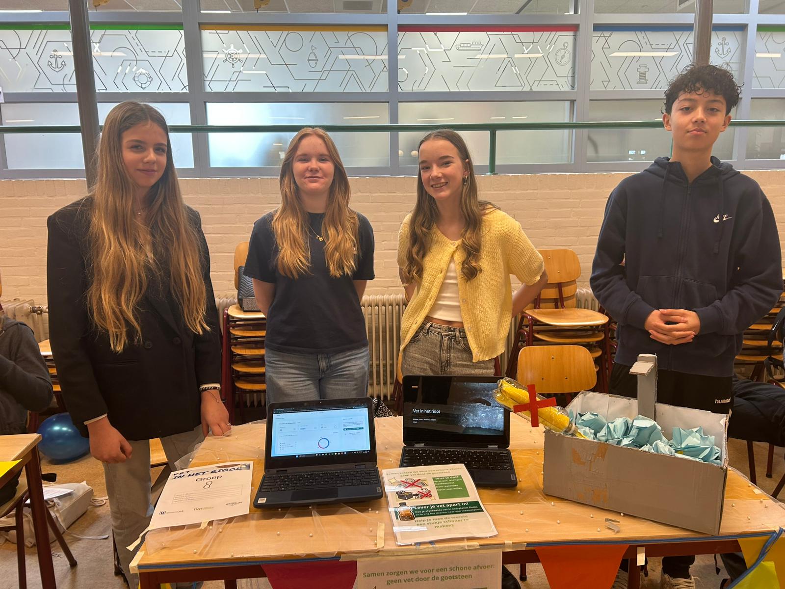 Vier studenten staan achter een tafel met laptops en een kartonnen project in een klaslokaal.