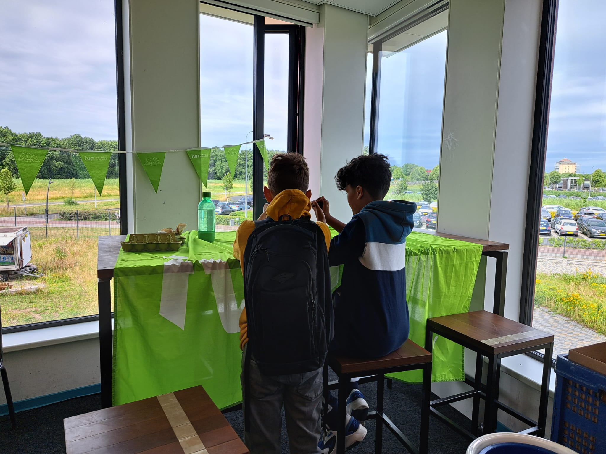 Twee kinderen zitten aan een tafel met groene decoratie en kijken naar buiten door een raam.