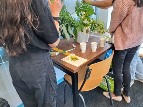 Twee mensen werken met bloemen op een tafel, omringd door bekers en planten.
