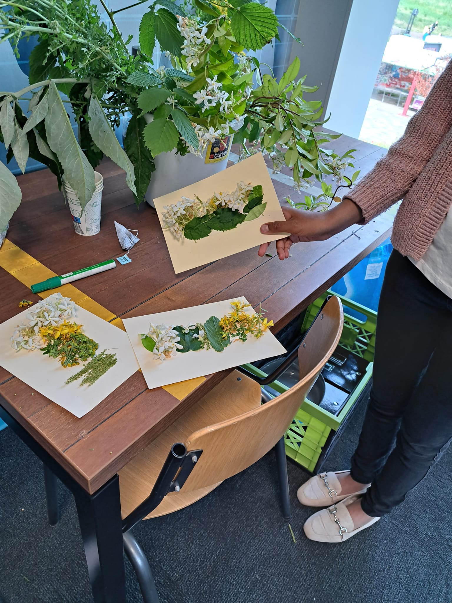 Persoon toont kaarten met gedroogde bloemen en bladeren op een tafel met groene planten.
