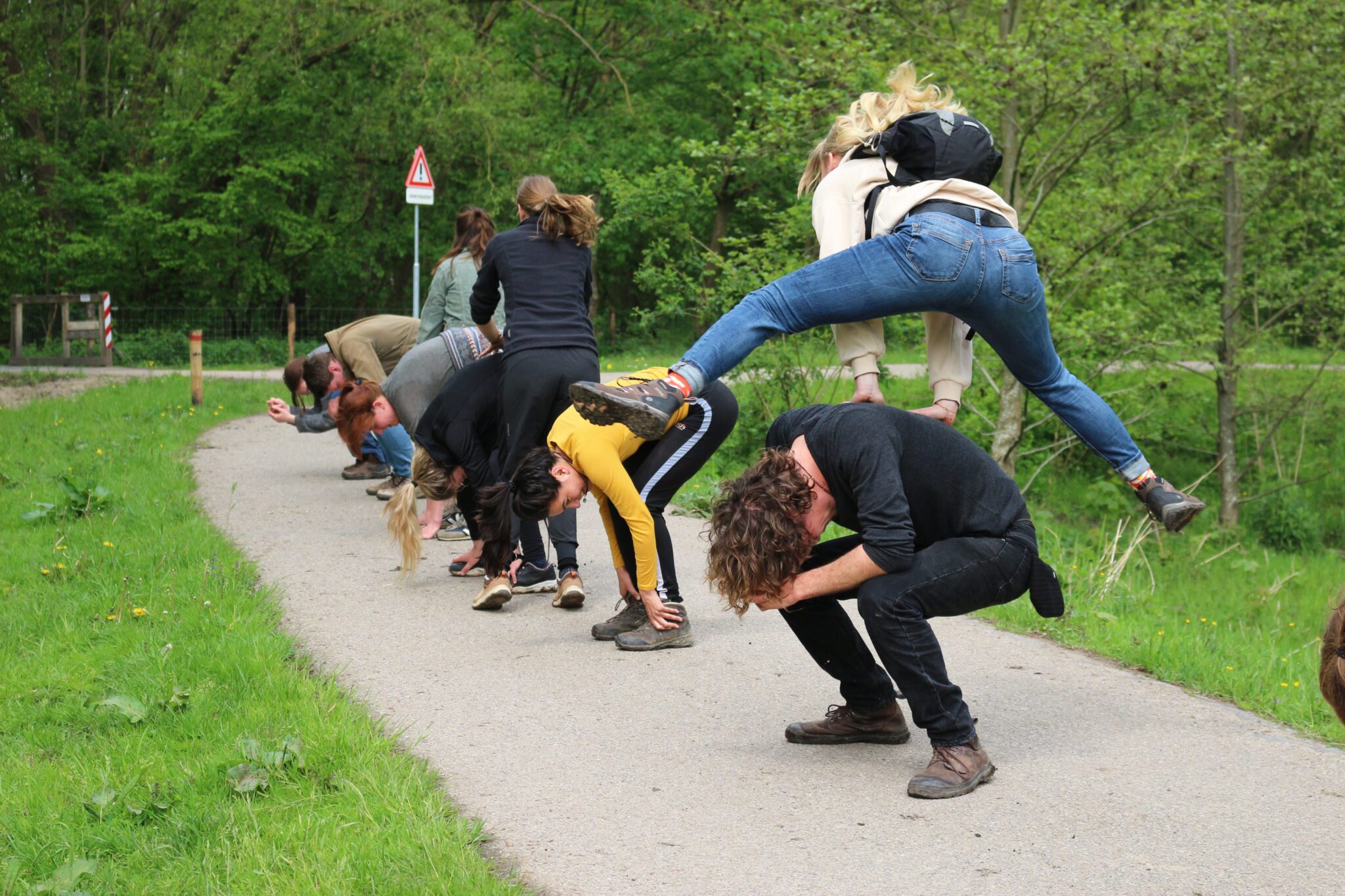 Personen doen een polonaise over gebogen deelnemers op een landweggetje bij een groene omgeving.
