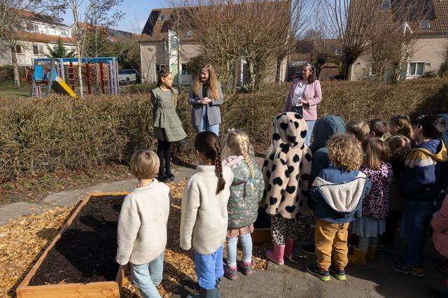 Kinderen luisteren naar volwassenen bij een bak met aarde; speeltuin en huizen in de achtergrond.