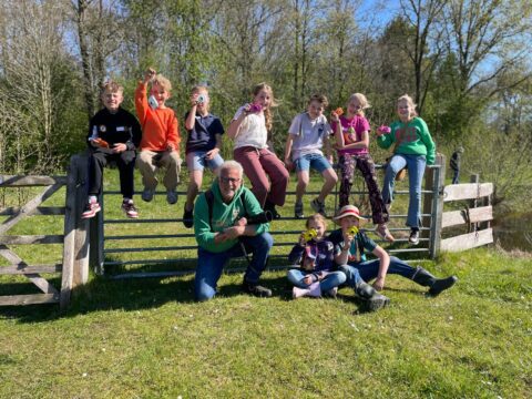 Groep kinderen en een volwassene, buiten op een hek, zonnige dag, groen gras en bomen op achtergrond.