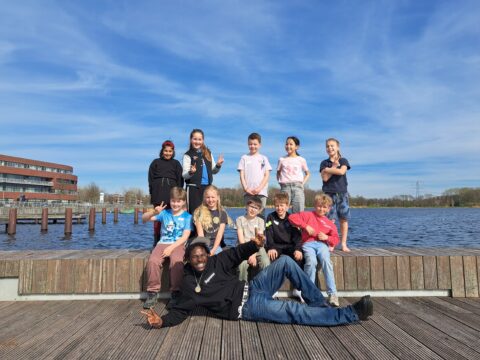 Groep kinderen poseert lachend op een steiger bij een meer, met een gebouw en een blauwe lucht op de achtergrond.