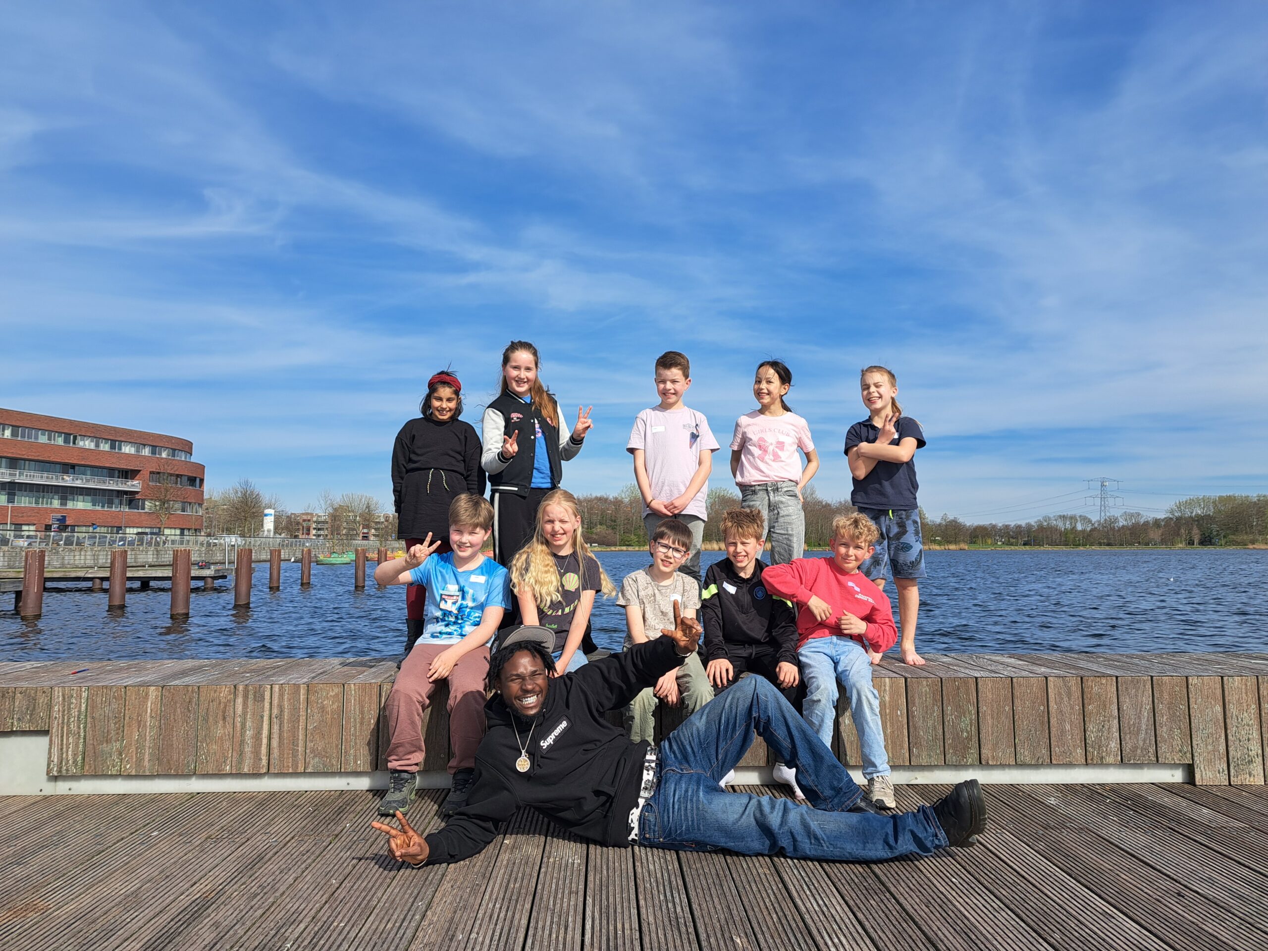 Groep kinderen poseert lachend op een steiger bij een meer, met een gebouw en een blauwe lucht op de achtergrond.