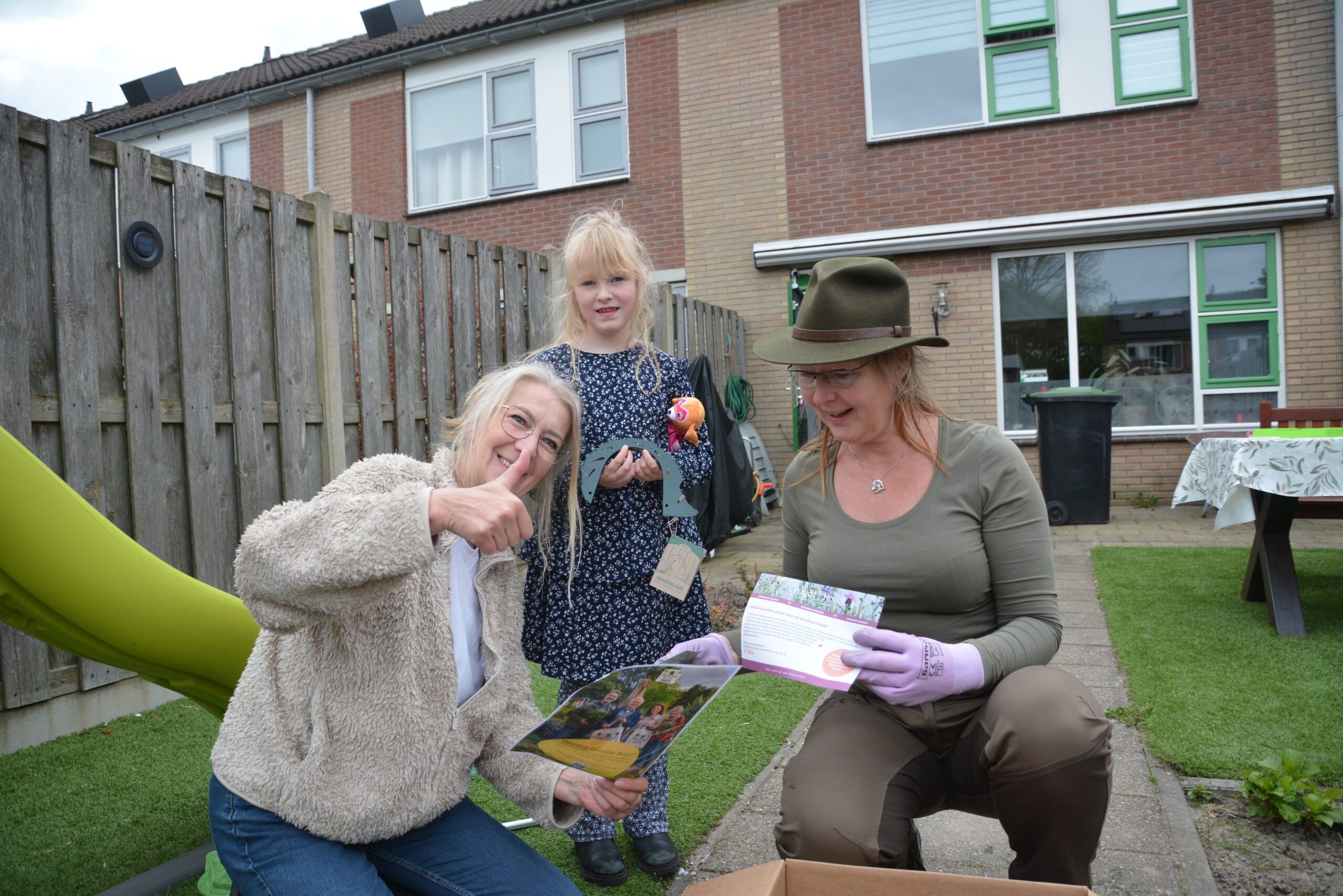 Twee vrouwen en een meisje openen een doos in de tuin. Een vrouw steekt duim op.