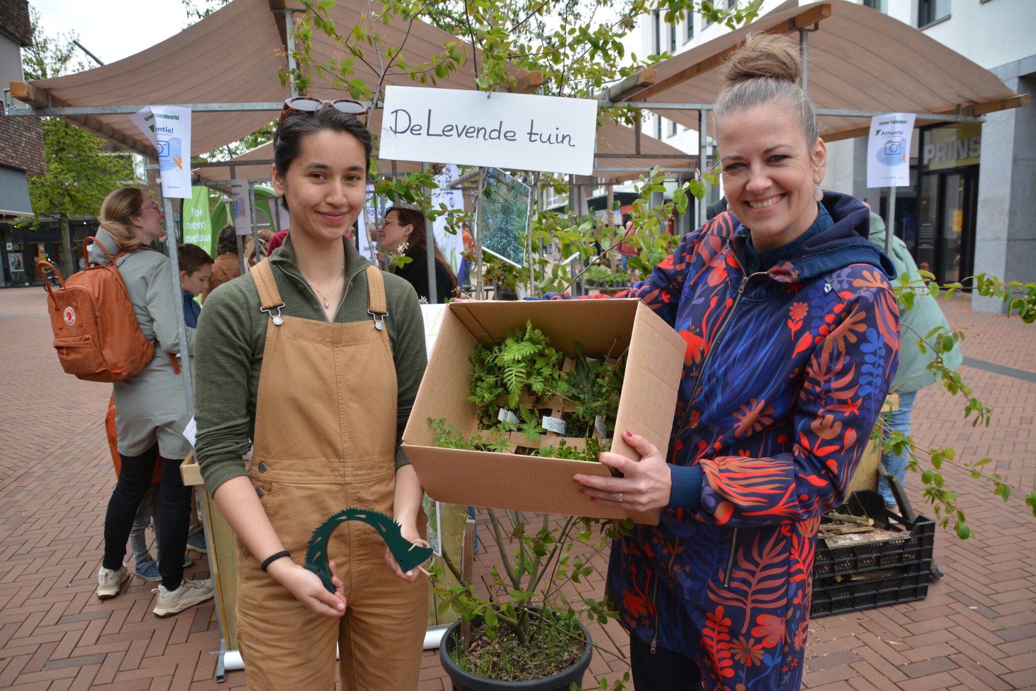 Twee vrouwen houden een doos met planten vast bij een kraam met het bord "De Levende tuin".