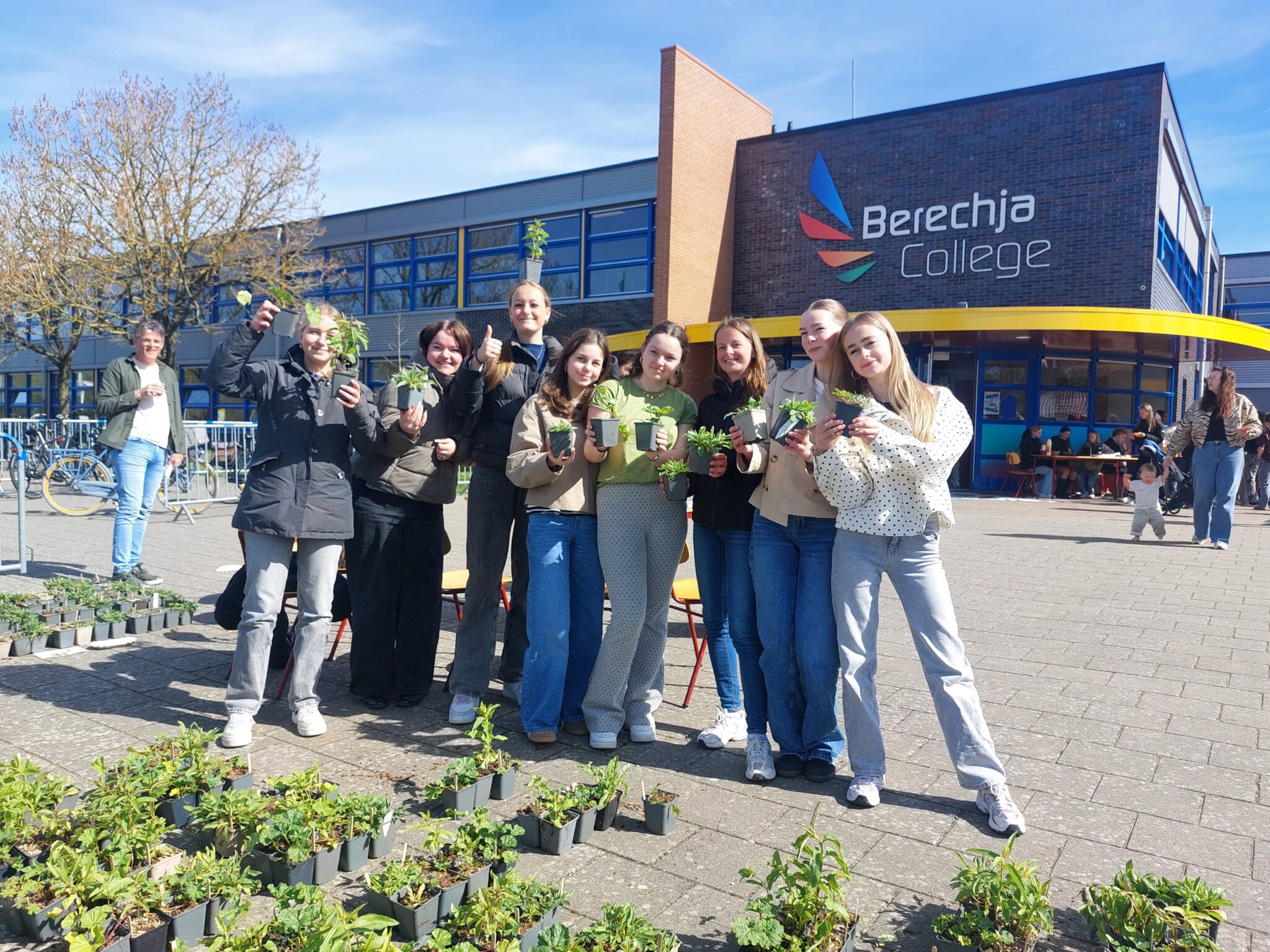 Groep jongeren met planten bij Berechja College op zonnige dag, met gebouw en fietsen op achtergrond.