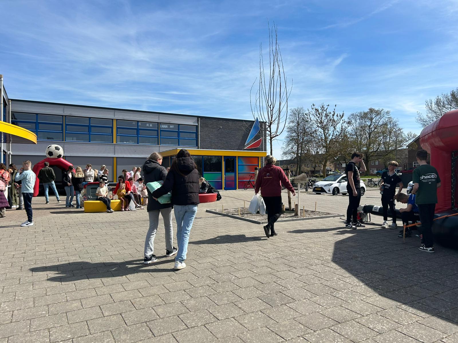 Mensen op schoolplein met opblaasbare structuur, kinderen zitten en staan, zonnige dag.