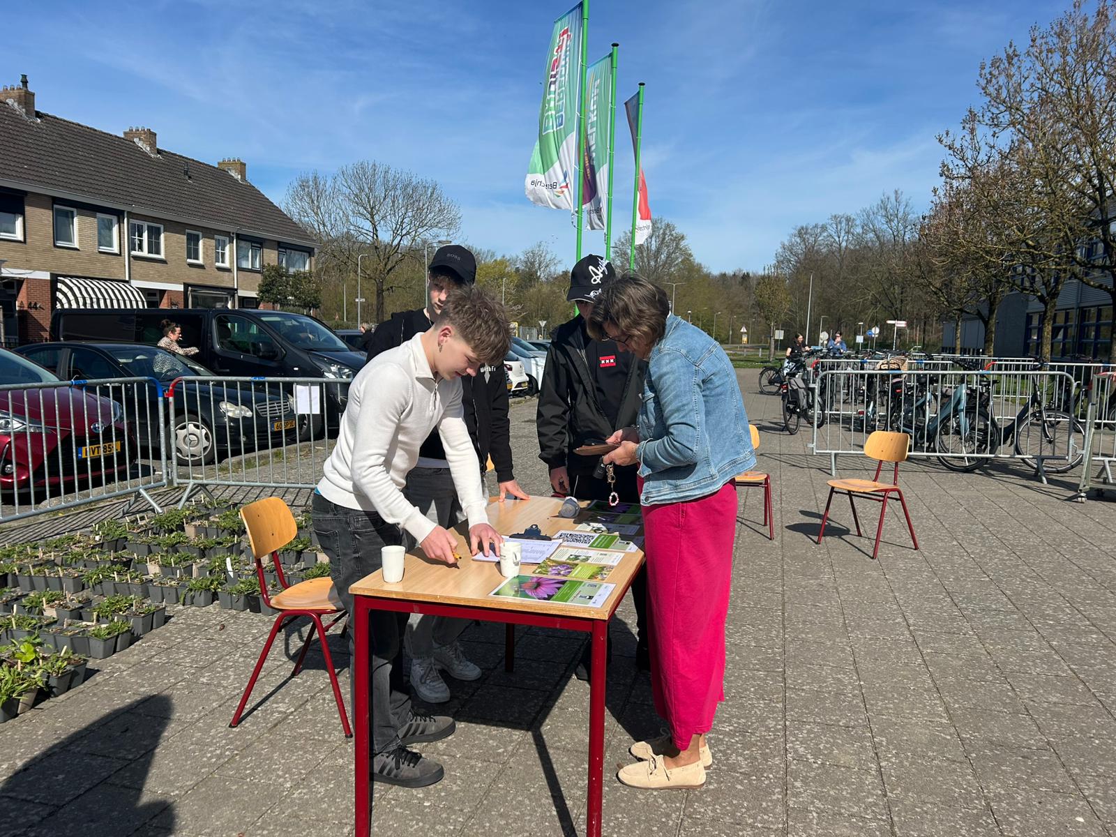 Personen aan tafel buiten met planten en papieren; fietsen en huizen op achtergrond.