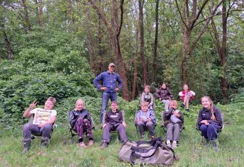 Groep kinderambassadeurs van de natuur en Michel Boeijen zitten en genieten van de natuur in een bosrijke omgeving.